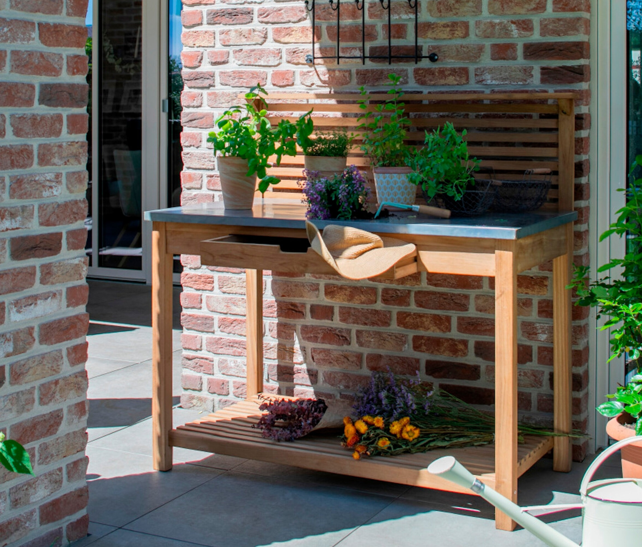 Ein deVries Teak-Pflanztisch mit Pflanzen und Gartenutensilien steht auf einer Terrasse.