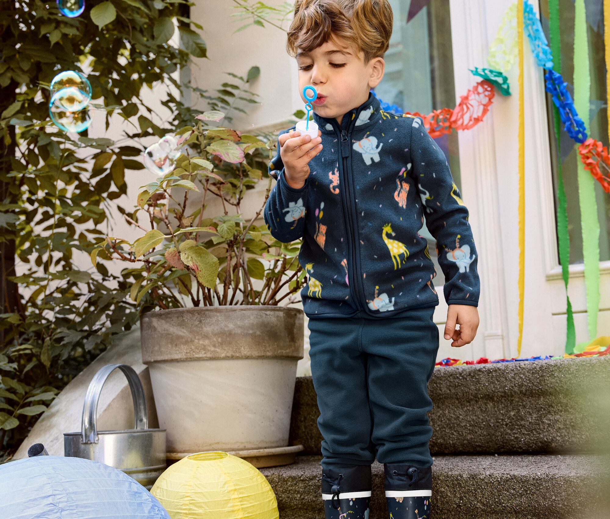 Junge pustet Blasen, trägt eine Kinder-Fleecejacke, Fleecehose und Gummistiefel.