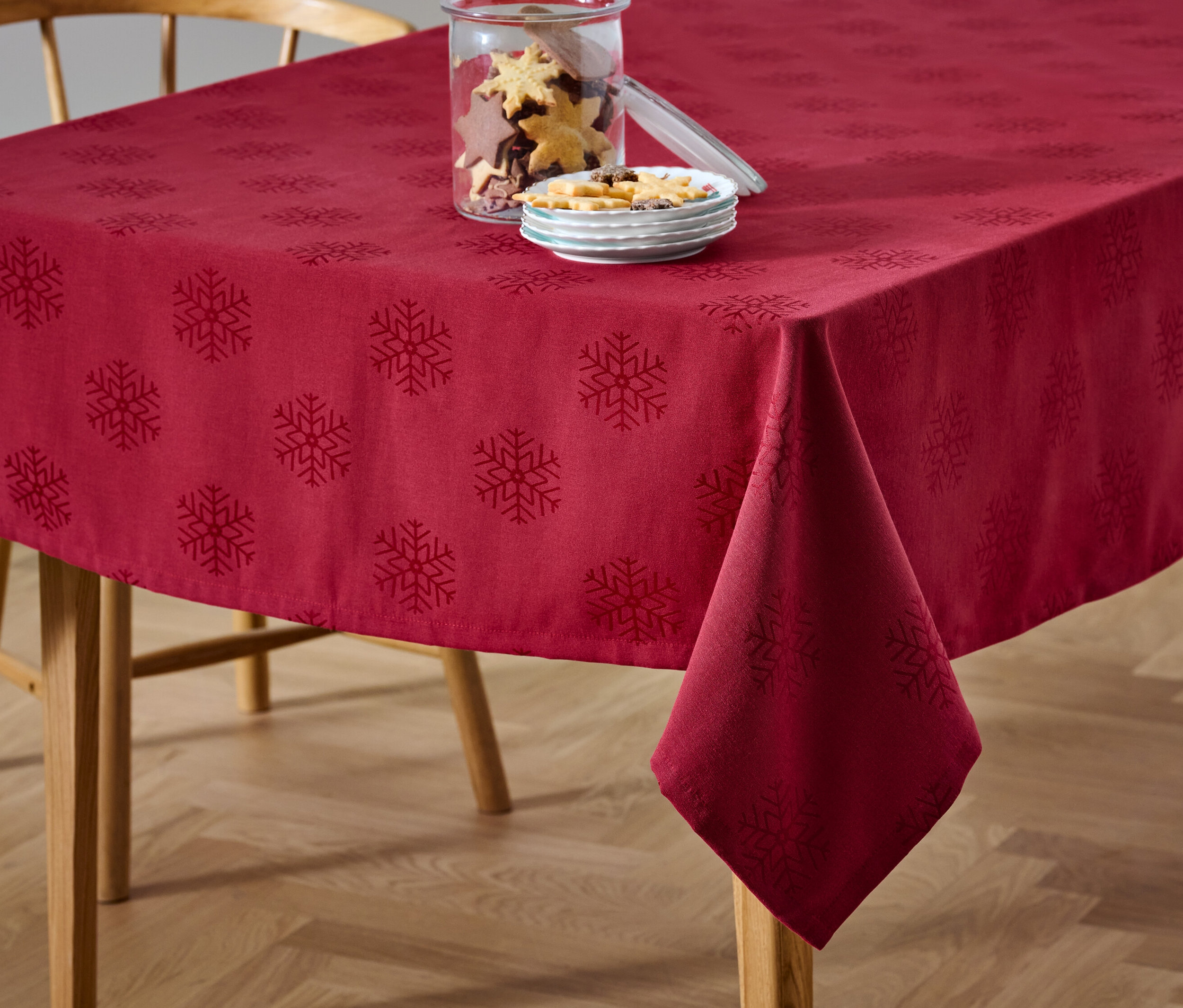 Eine rote Jacquard-Tischdecke mit Schneeflockenmuster liegt auf einem Tisch neben einem Glas mit Keksen und einem Teller mit Süßigkeiten.