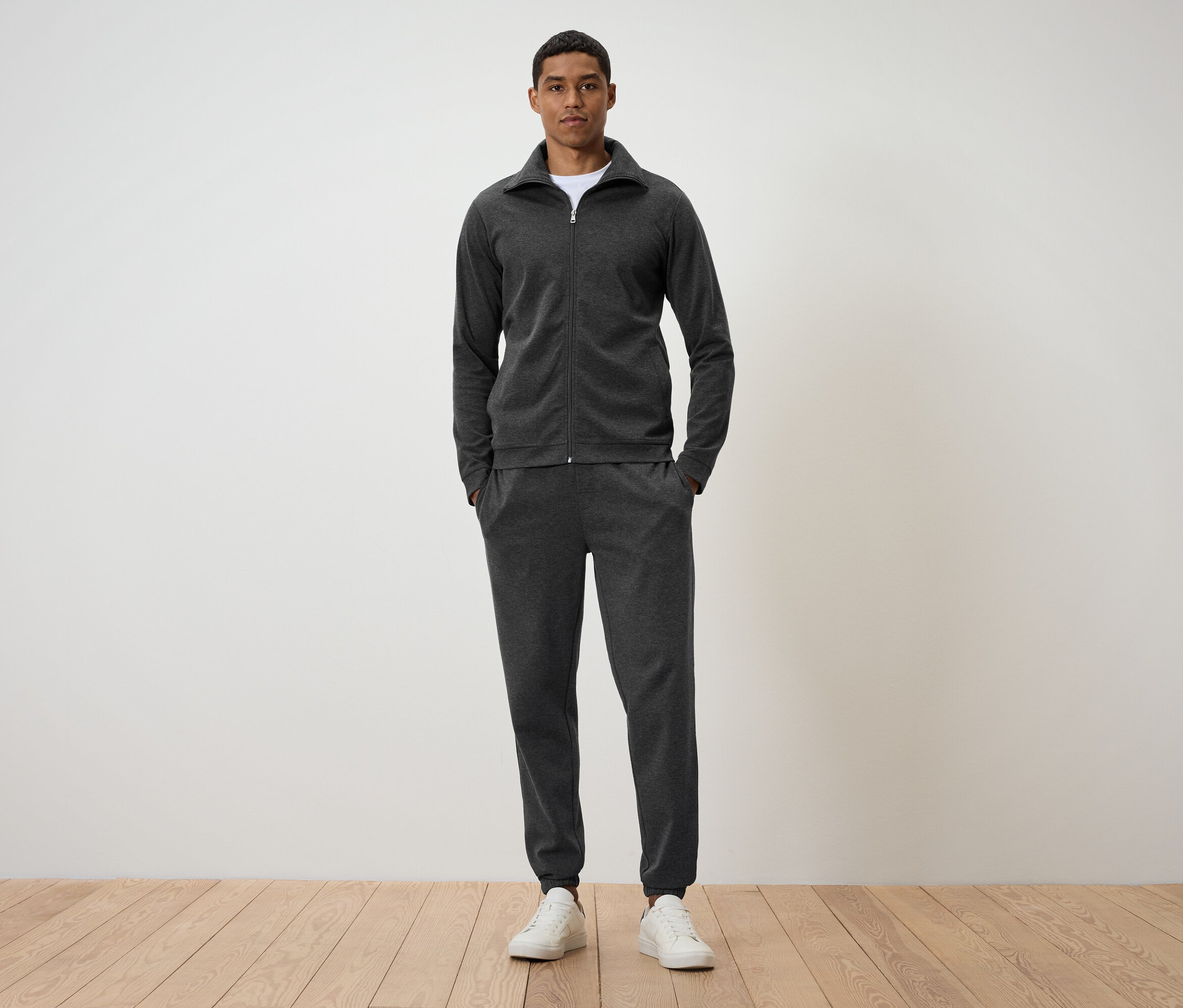 Ein Mann steht auf einem Holzboden und trägt einen dunkelgrauen Trainingsanzug und weiße Turnschuhe.