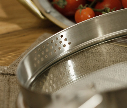 Detail eines Glasdeckels mit Edelstahlrand mit Dampflöchern und roten Tomaten im Hintergrund.