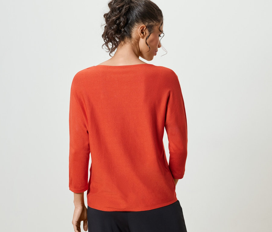 Rückansicht einer Frau mit hochgesteckten Haaren, die einen orangefarbenen Feinstrickpullover und eine schwarze Hose trägt.