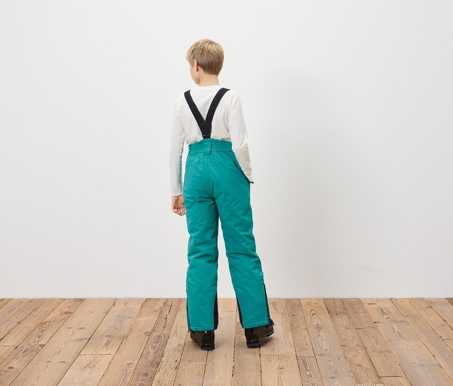 Ein Junge steht mit dem Rücken zur Kamera und trägt eine türkisfarbene Skihose mit schwarzen Hosenträgern.