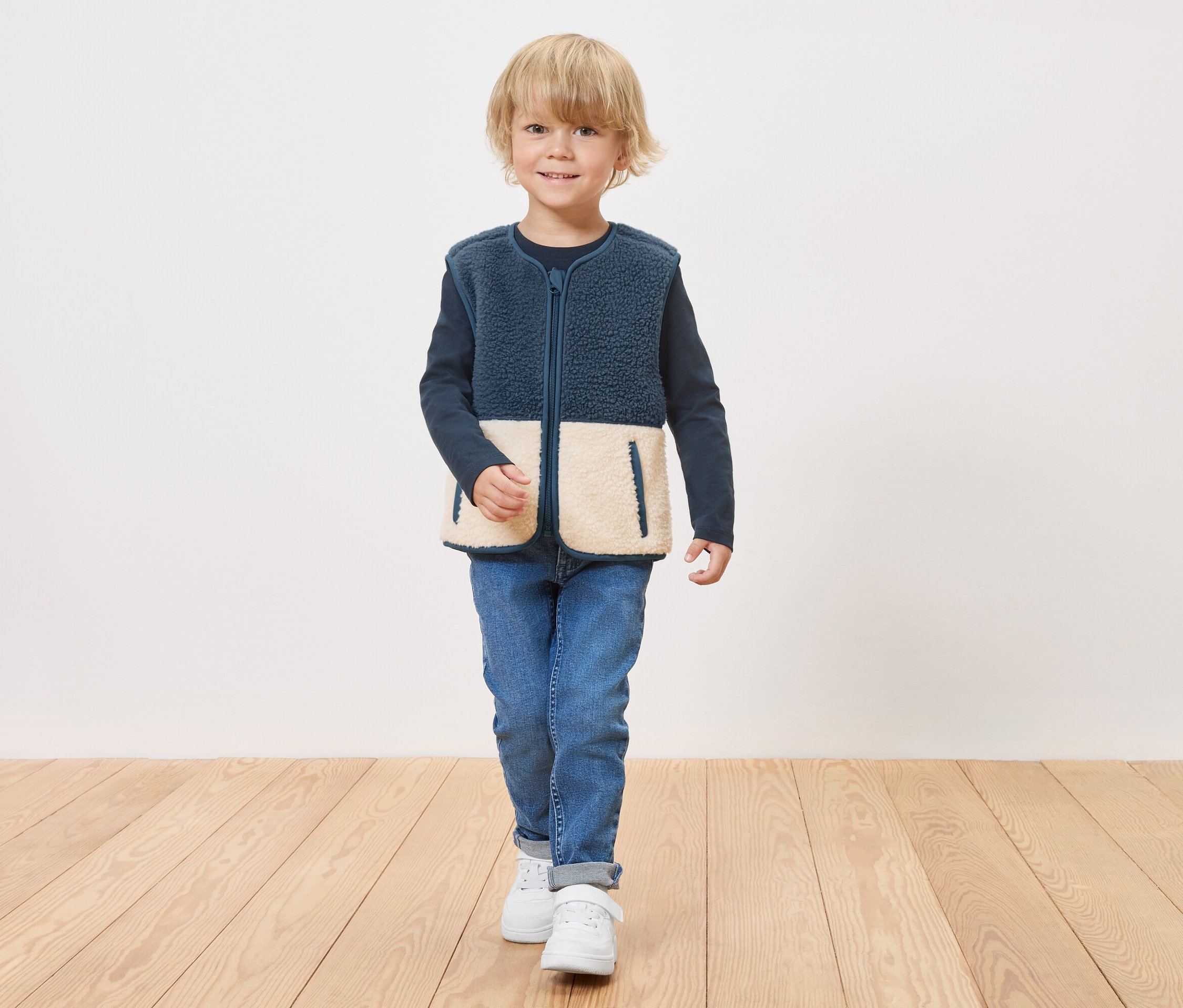 Ein Junge in einer marineblauen Fleeceweste mit cremefarbenen unteren Einsätzen mit Reißverschluss steht auf einem Holzboden.