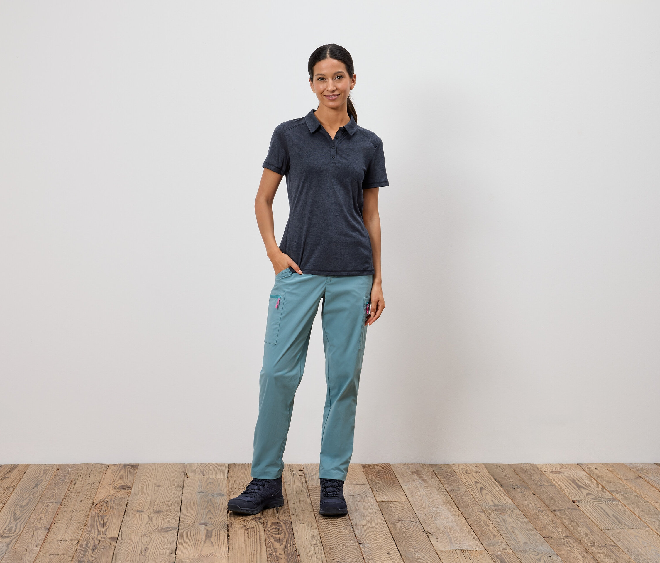 Eine Frau steht mit einer Hand in der Tasche vor einer weißen Wand.