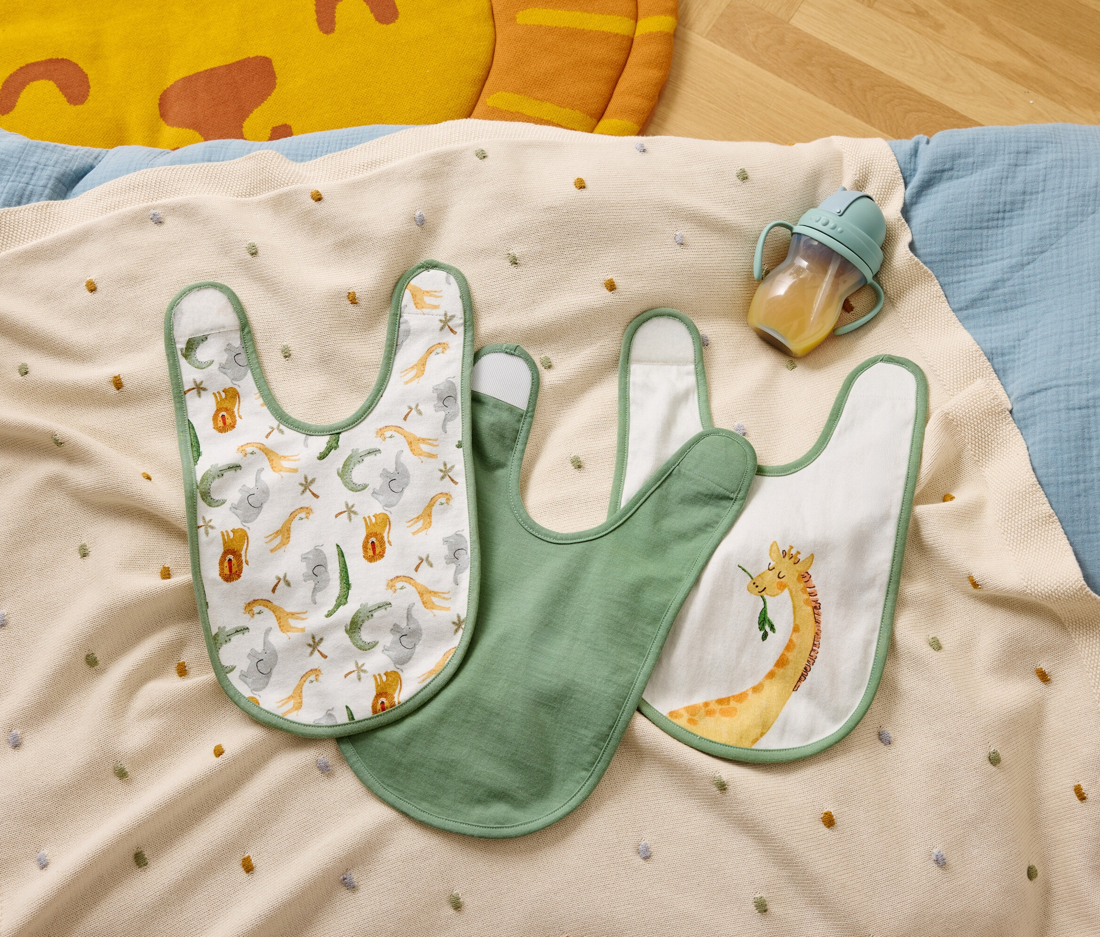 Drei Lätzchen und eine Trinkflasche liegen auf einer beigen Decke mit bunten Punkten.