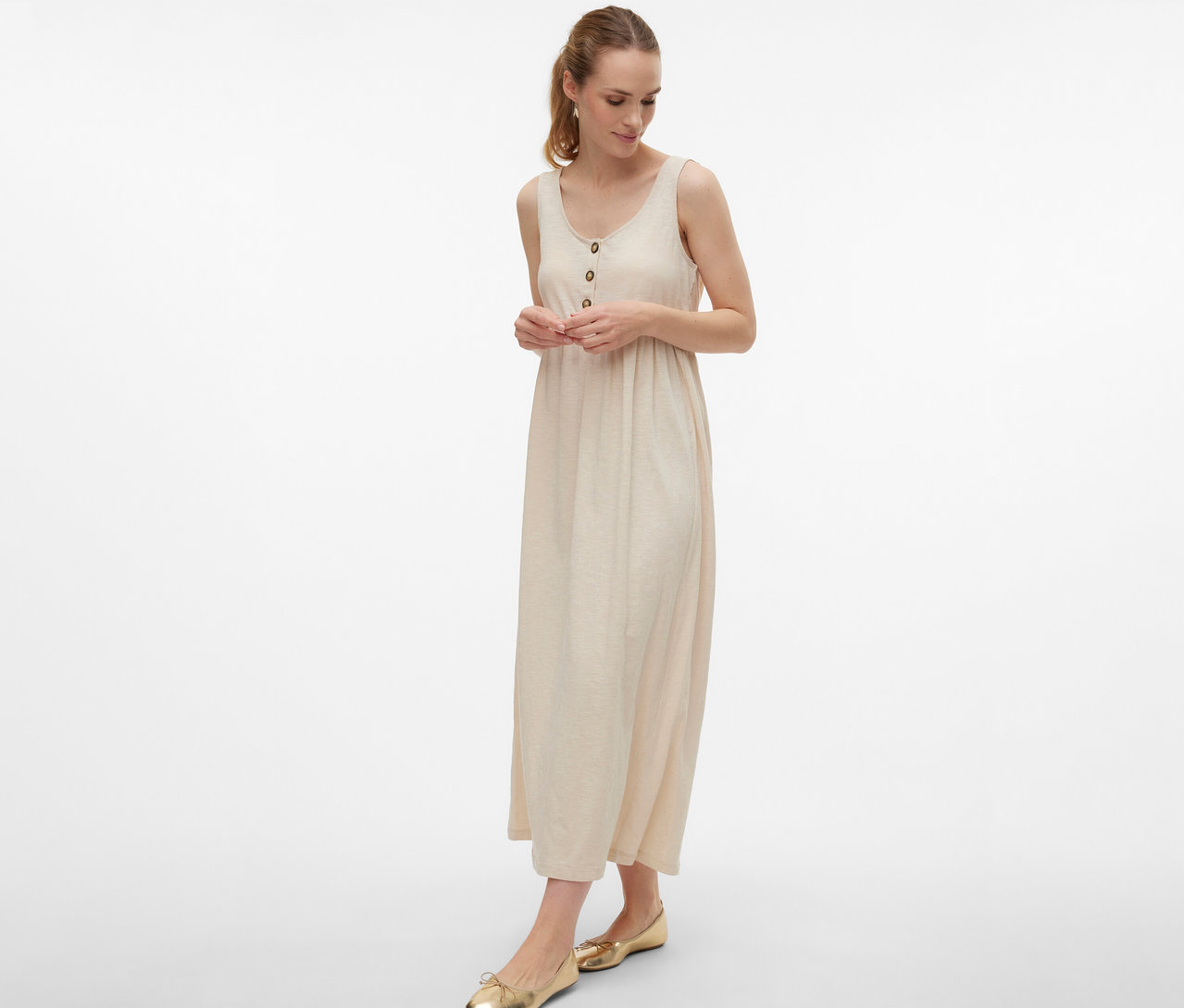 Frau trägt MAMALICIOUS Schwangerschaftskleid »MLEVI LIA« mit Knöpfen und goldene Ballerinas vor weißem Hintergrund.