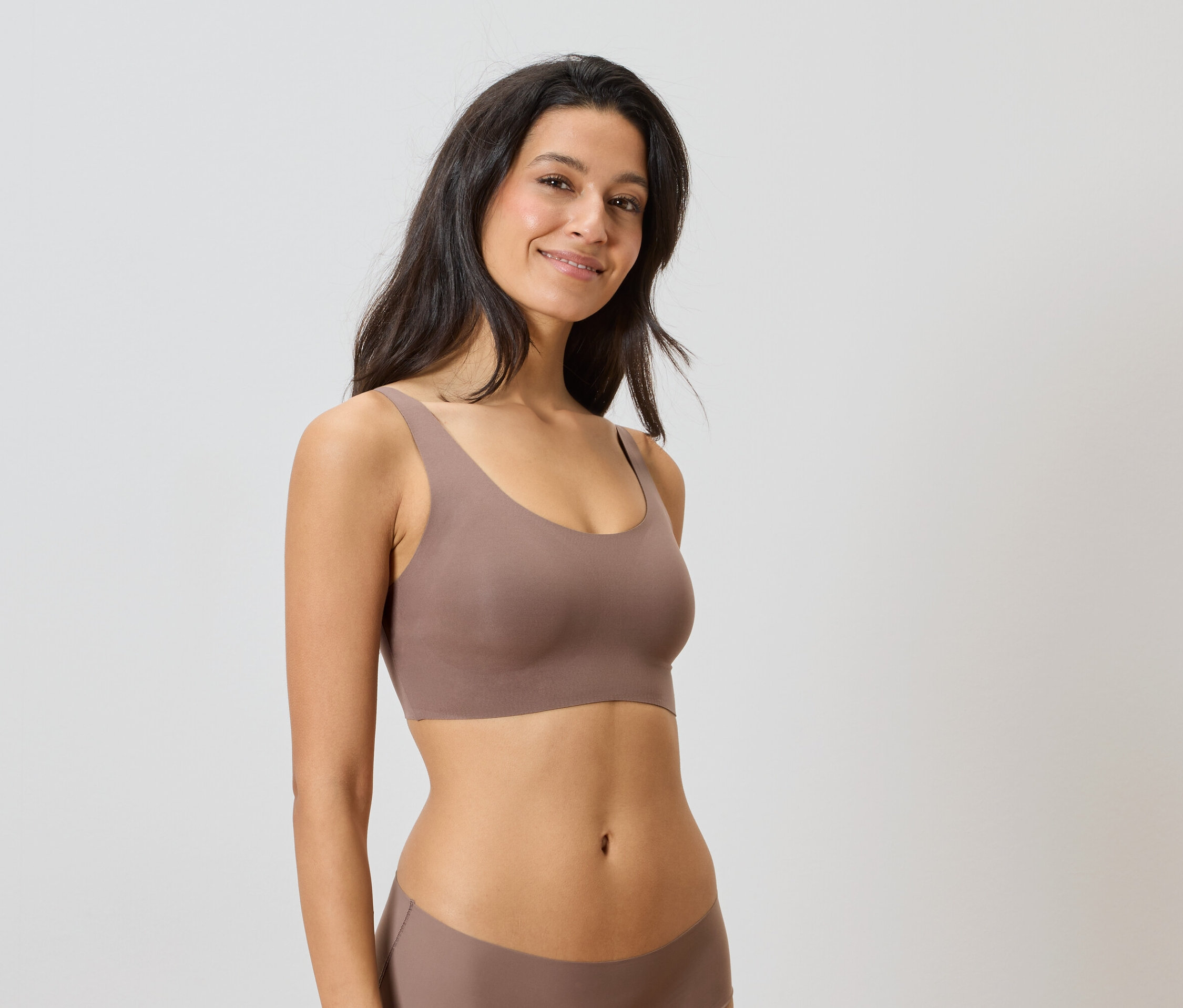 Frau trägt ein braunes Unterwäsche-Set vor einem neutralen Hintergrund.