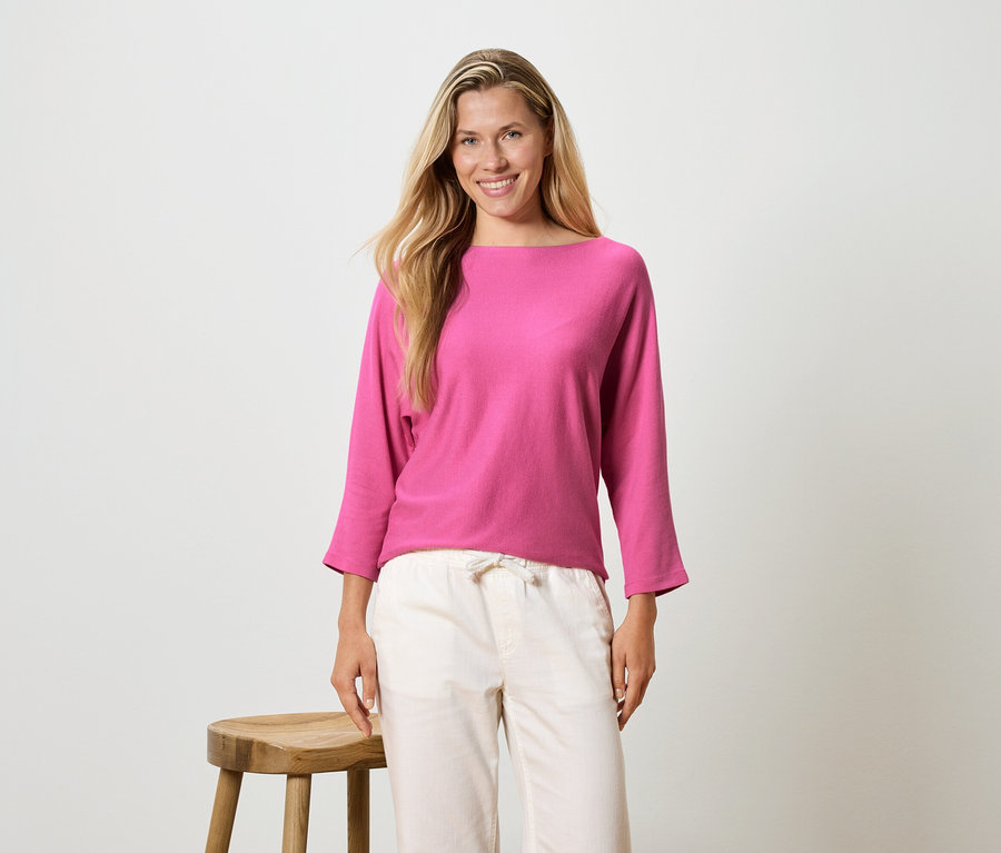 Frau mit langen blonden Haaren trägt ein pinkfarbenes Langarmshirt und eine helle Hose und steht neben einem Hocker.