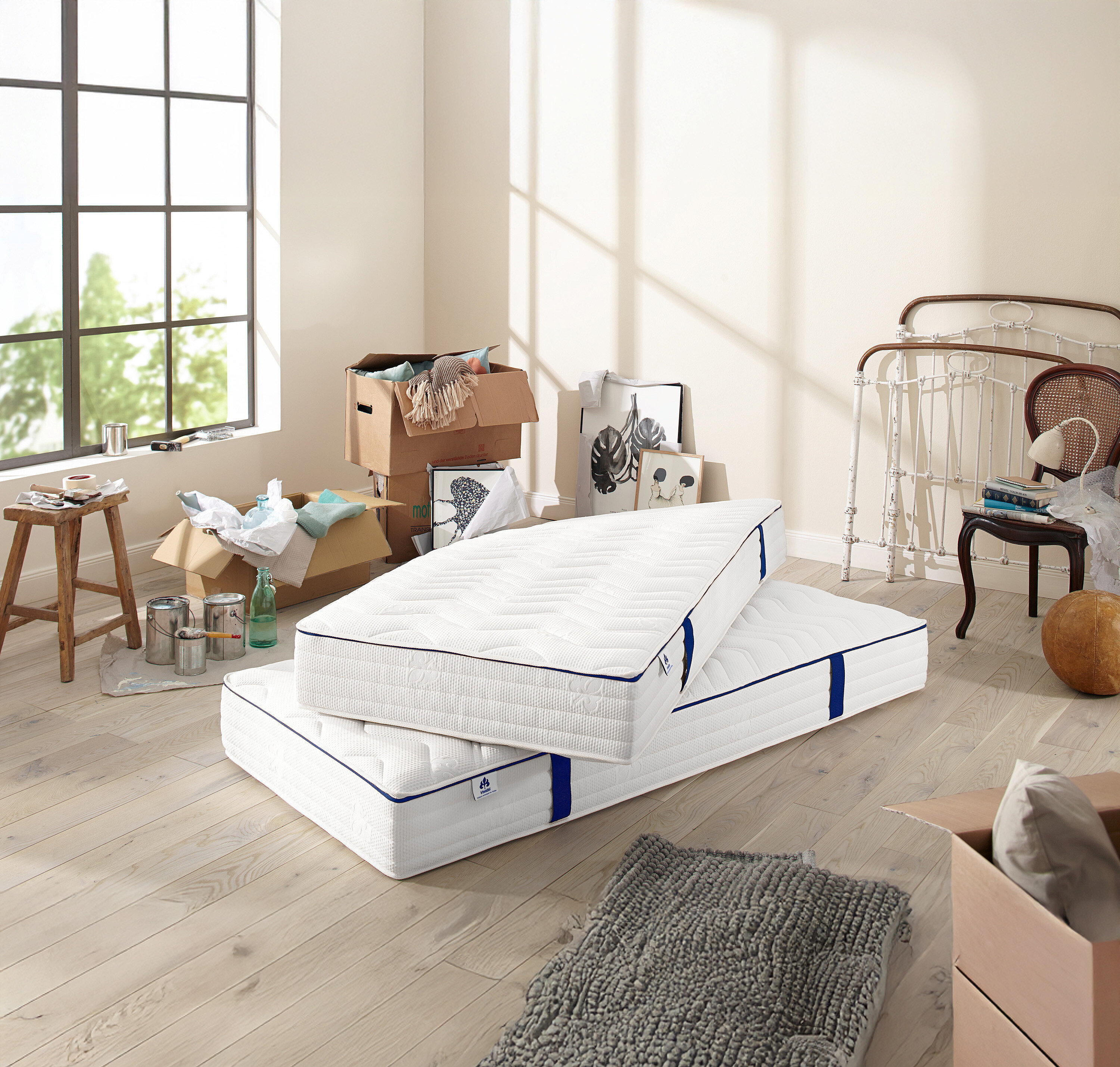 Zwei weiße Matratzen mit blauer Bordüre liegen übereinander in einem Zimmer. Um sie herum stehen Kisten mit Sachen.