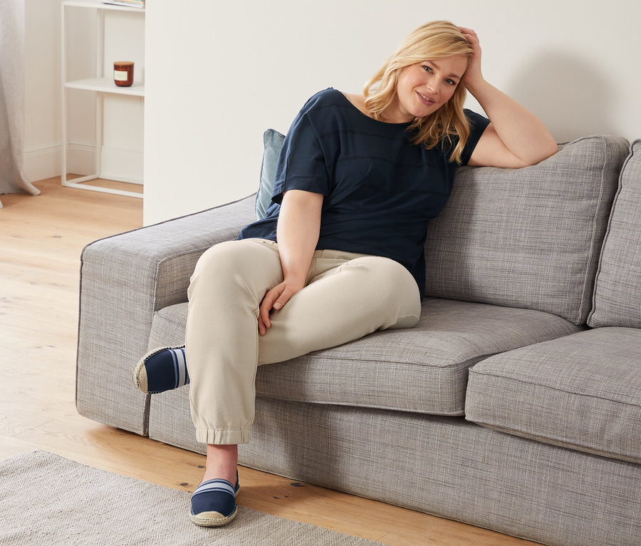 Blonde Frau sitzt auf einem grauen Sofa, trägt ein dunkelblaues Shirt mit Rundhalsausschnitt, knöchellange Joggpants und Espadrilles.