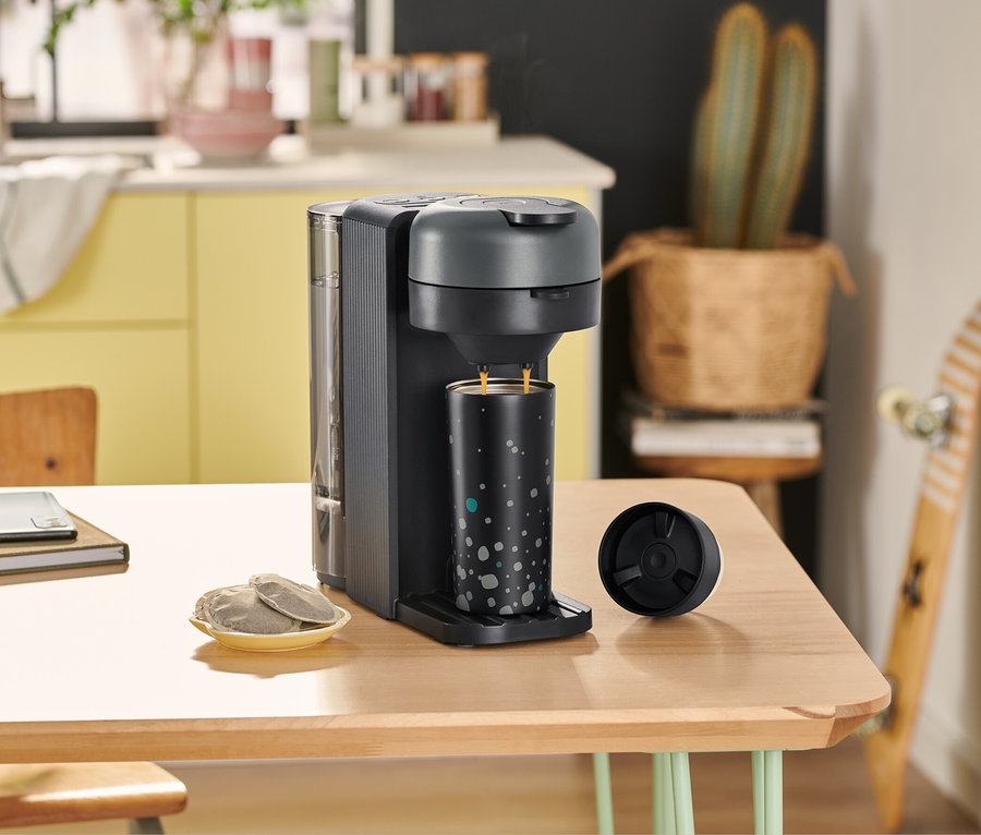 Auf einem Tisch steht eine »Call me Pad« Kaffeepadmaschine in Graphit, eine Tasse, ein Glas Wasser und ein Notizbuch.