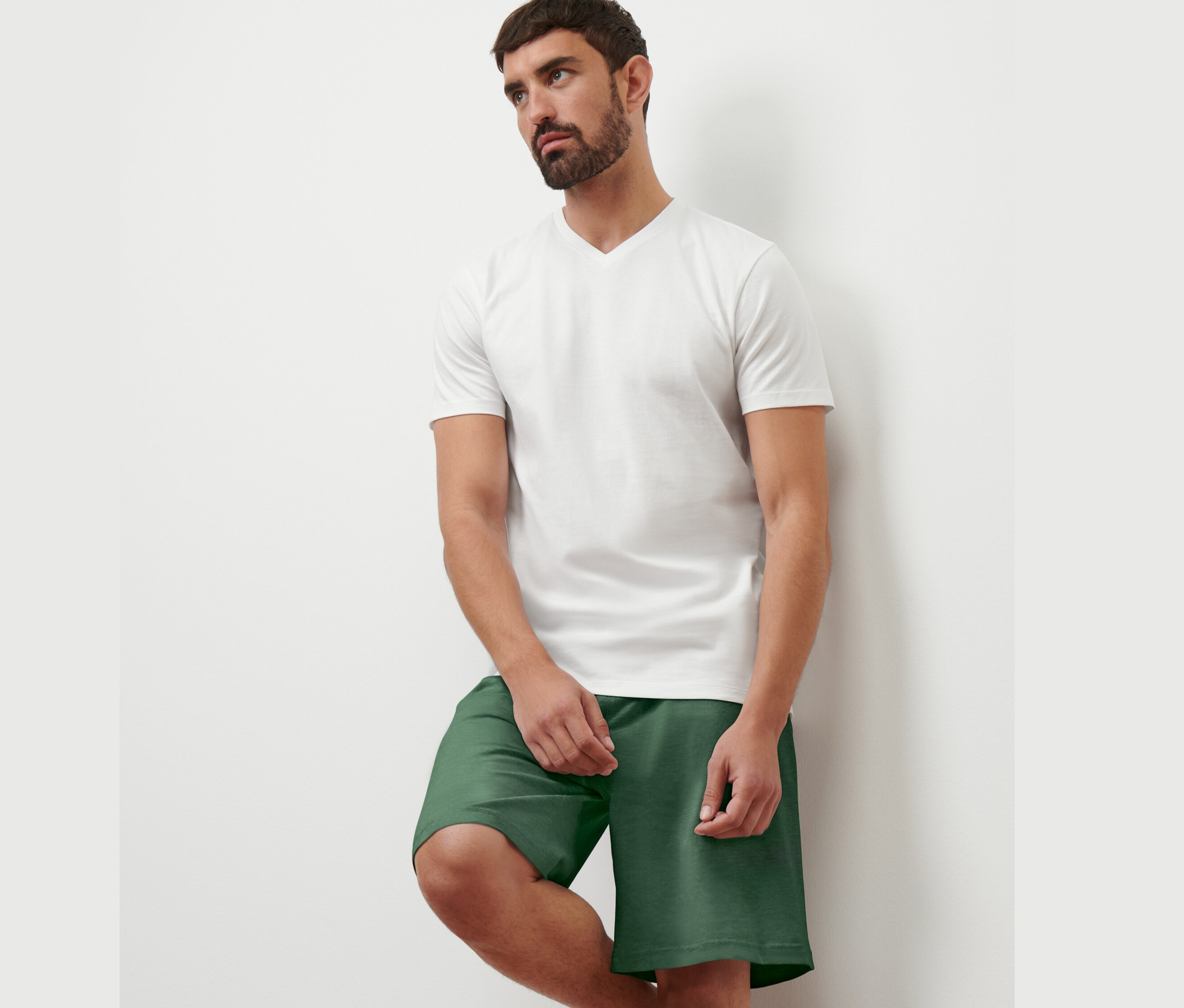 Mann lehnt an einer Wand, trägt ein weißes T-Shirt mit V-Ausschnitt und grüne Shorts. Er trägt 1 von 2 T-Shirts mit V-Ausschnitt.