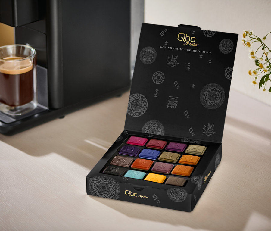 Eine schwarze Qbo ICONIC Dark Chrome Kaffeemaschine mit einem Glas Kaffee auf der Abtropfschale steht neben einer geöffneten schwarzen Box Qbo Collection - 16 Kapseln, die 16 bunte Kaffeepads zeigt. Im Hintergrund steht eine weiße Vase mit weißen und gelben Blumen.