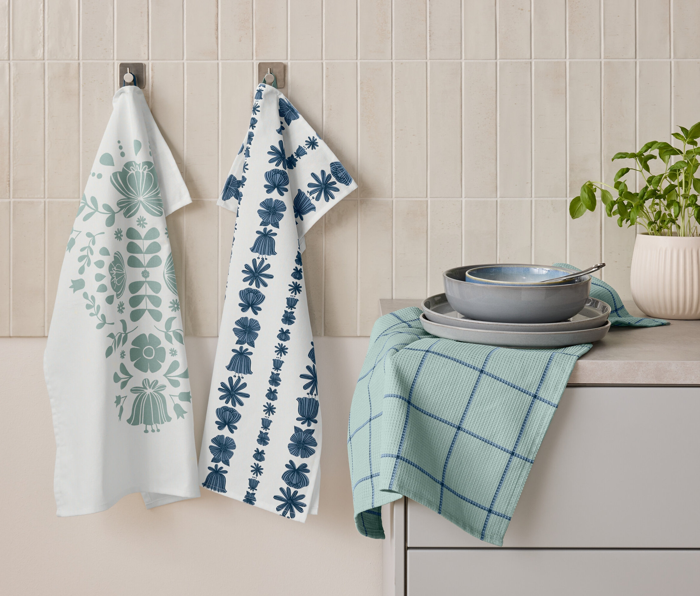 Zwei Geschirrtücher mit floralen Mustern hängen nebeneinander an der Wand. Darunter liegt ein gefaltetes grünes Geschirrtuch mit blauen Streifen, auf dem Teller und eine Schüssel gestapelt sind. Daneben steht ein Topf mit Kräutern.