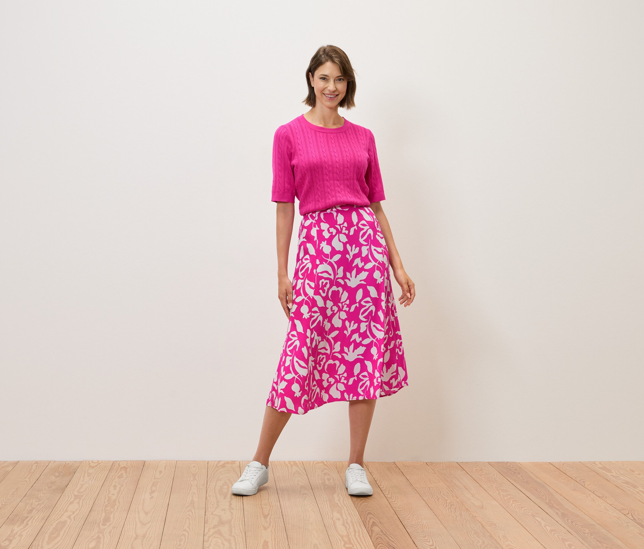 Frau steht, trägt ein pinkes Oberteil und einen Rock mit Blumenmuster.