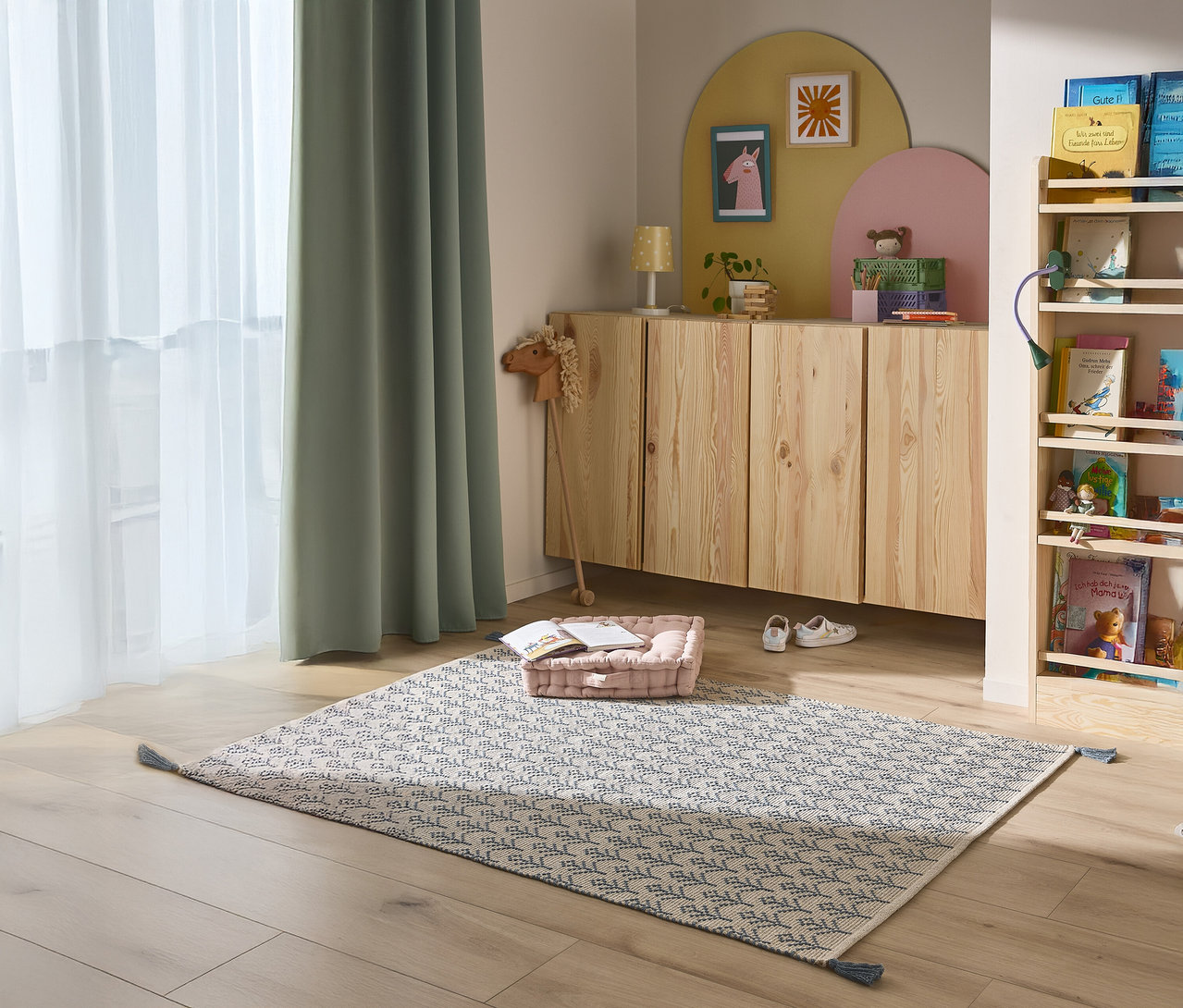 Ein Kinderzimmer mit einem gemusterten Teppich, einem niedrigen Schrank, einem Bücherregal und grünen Vorhängen.