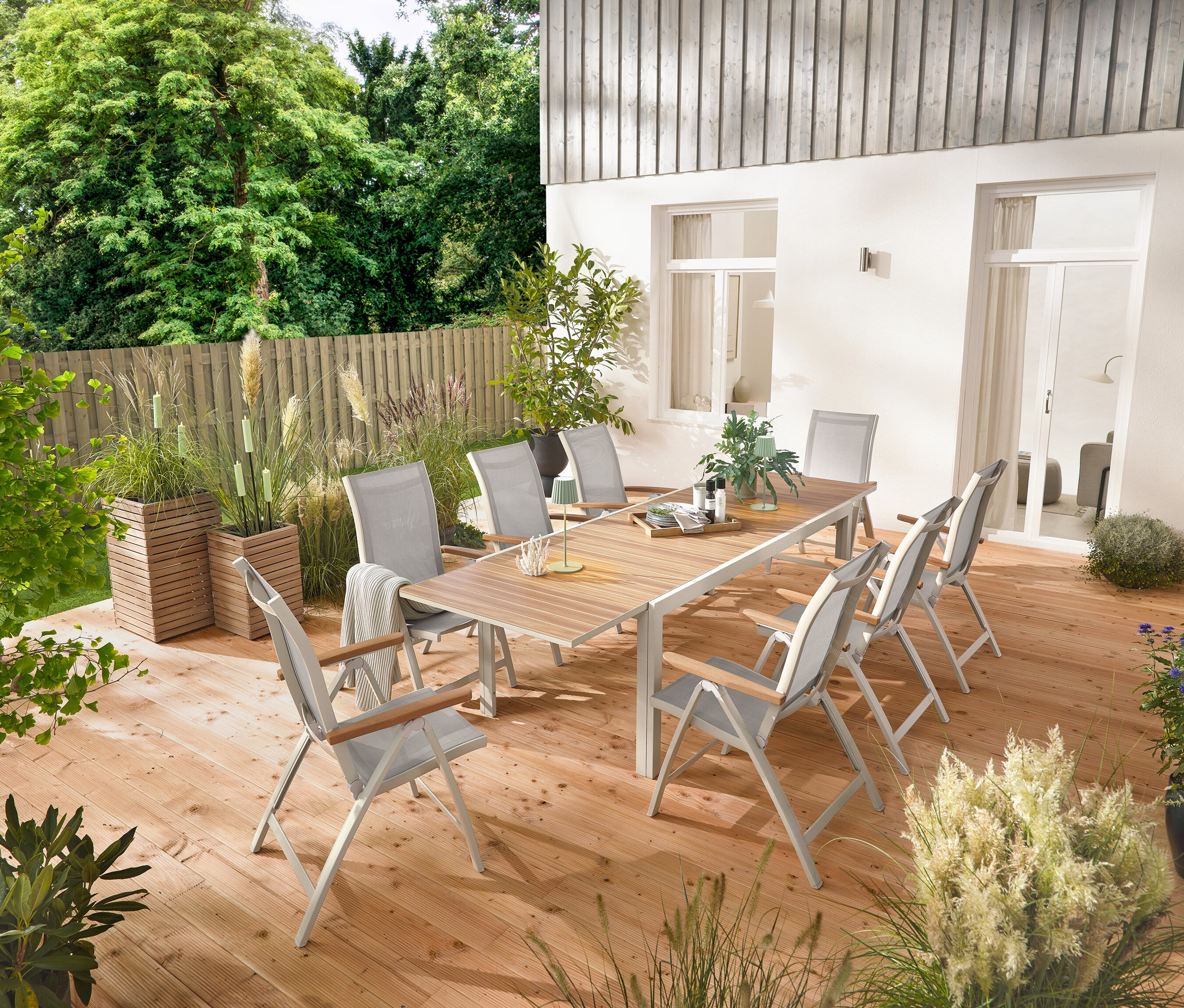 Ein ausziehbarer Gartentisch aus Aluminium »Liska« mit ca. 200–300 cm Länge steht auf einer Holzterrasse, umgeben von sechs Hochlehnern »Liska« mit verstellbarer Rückenlehne. Zwei Pflanzsäulen »Tinus« und fünf LED-Gartenfackeln sind ebenfalls sichtbar.