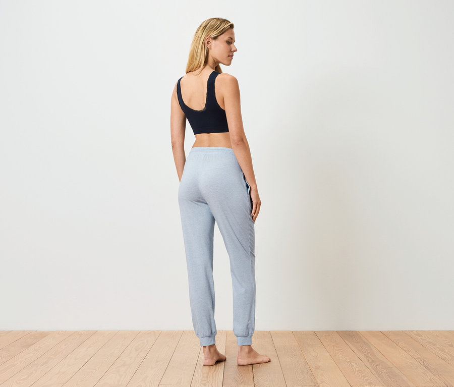 Frau steht mit dem Rücken zum Betrachter, trägt einen schwarzen Sport-BH und hellblaue Jogginghose.