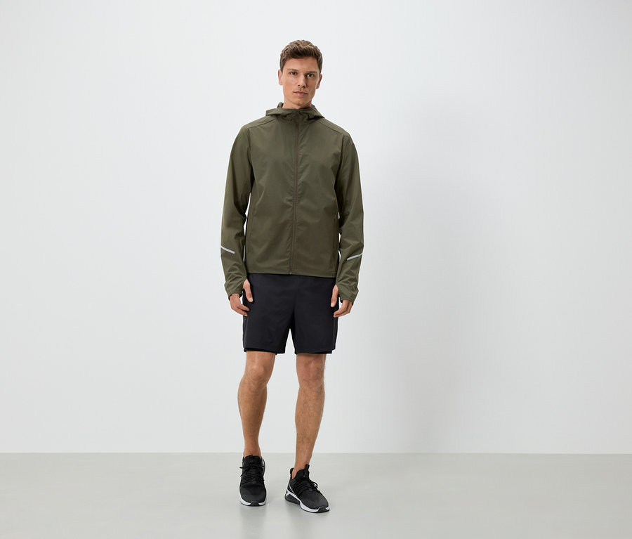 Mann in olivgrüner Jacke mit Kapuze, schwarzen Shorts und Turnschuhen.