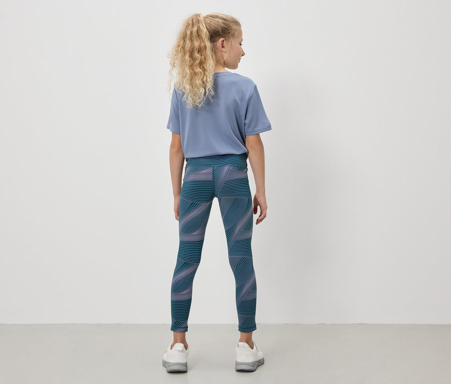 Mädchen mit blonden Haaren trägt ein blaues T-Shirt und Leggings mit geometrischem Muster.