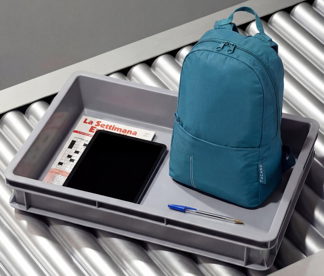 Ein petrolfarbener Tucano Rucksack »Compatto Eco« steht in einer grauen Wanne mit Tablet, Zeitschrift und Stift auf einem Rollband.