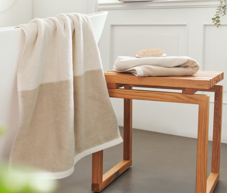 Ein cremeweißes Jacquard-Handtuch hängt über dem Badewannenrand, zwei hochwertige Handtücher liegen auf einem Holzschemel.