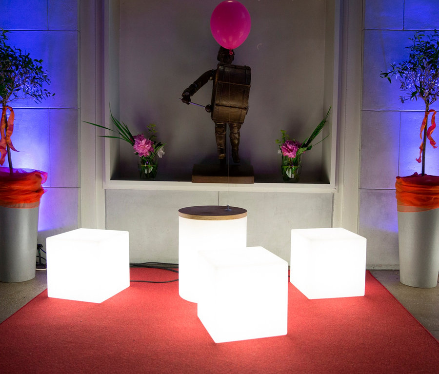 Beleuchtete Würfel und Pflanzgefäße säumen einen roten Teppich, im Hintergrund eine Trommlerstatue.