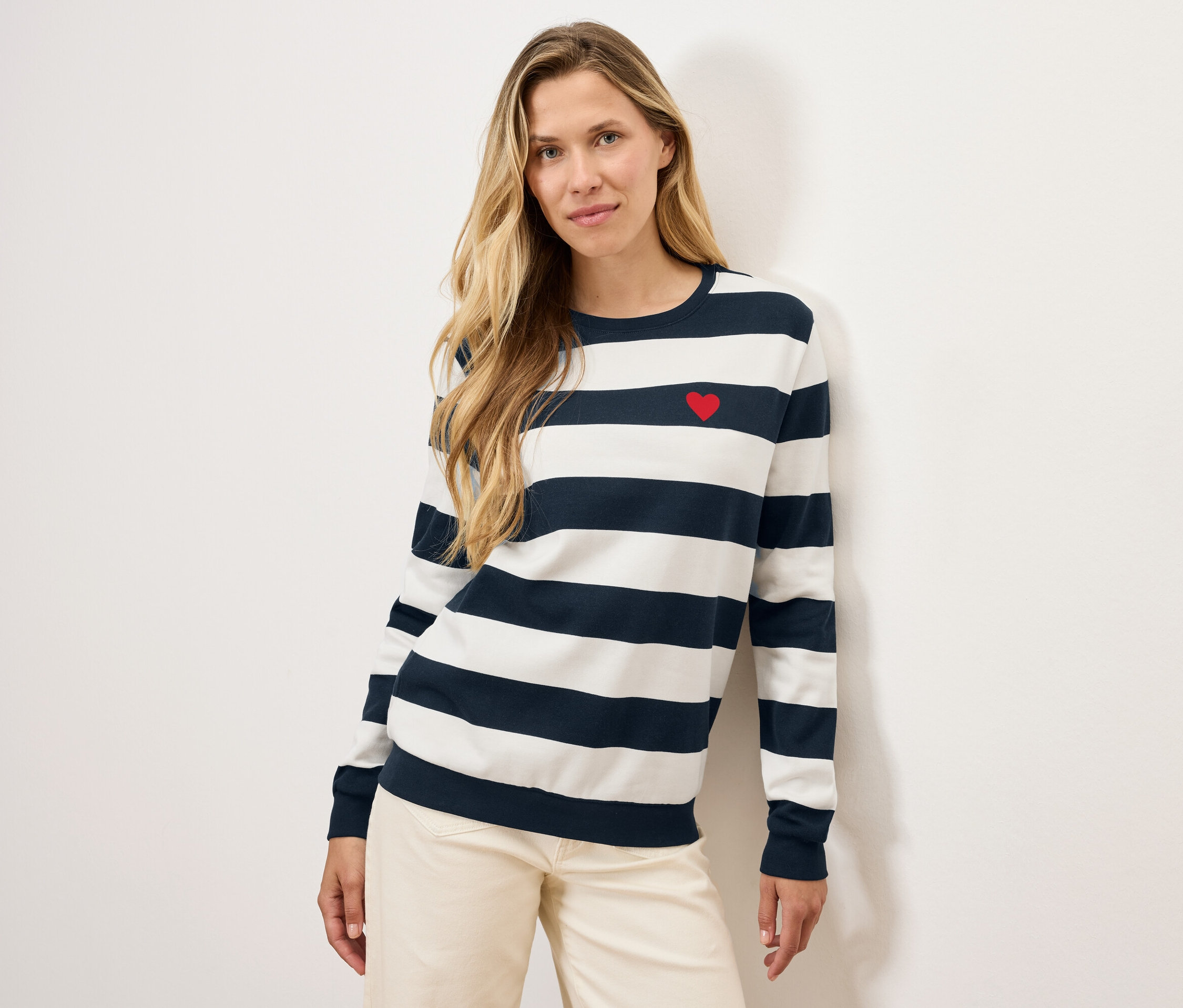 Blonde Frau posiert in einem gestreiften Pullover mit rotem Herz und heller Hose.