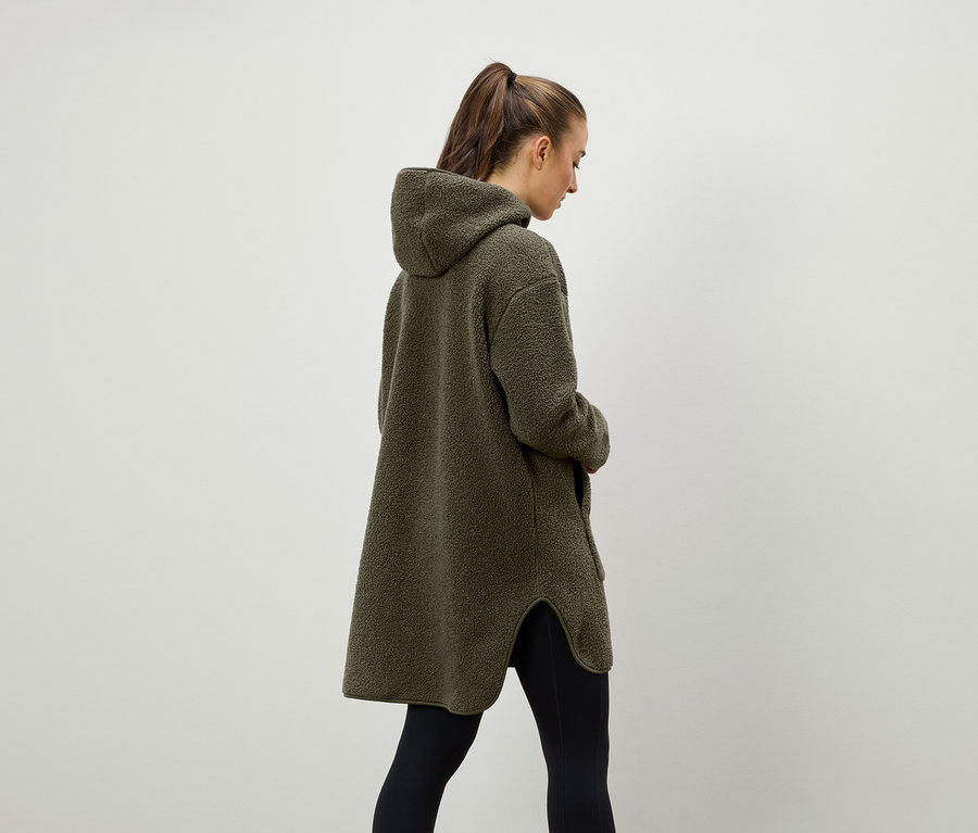 Eine Frau von hinten trägt einen olivgrünen, flauschigen Kapuzenmantel und schwarze Leggings.