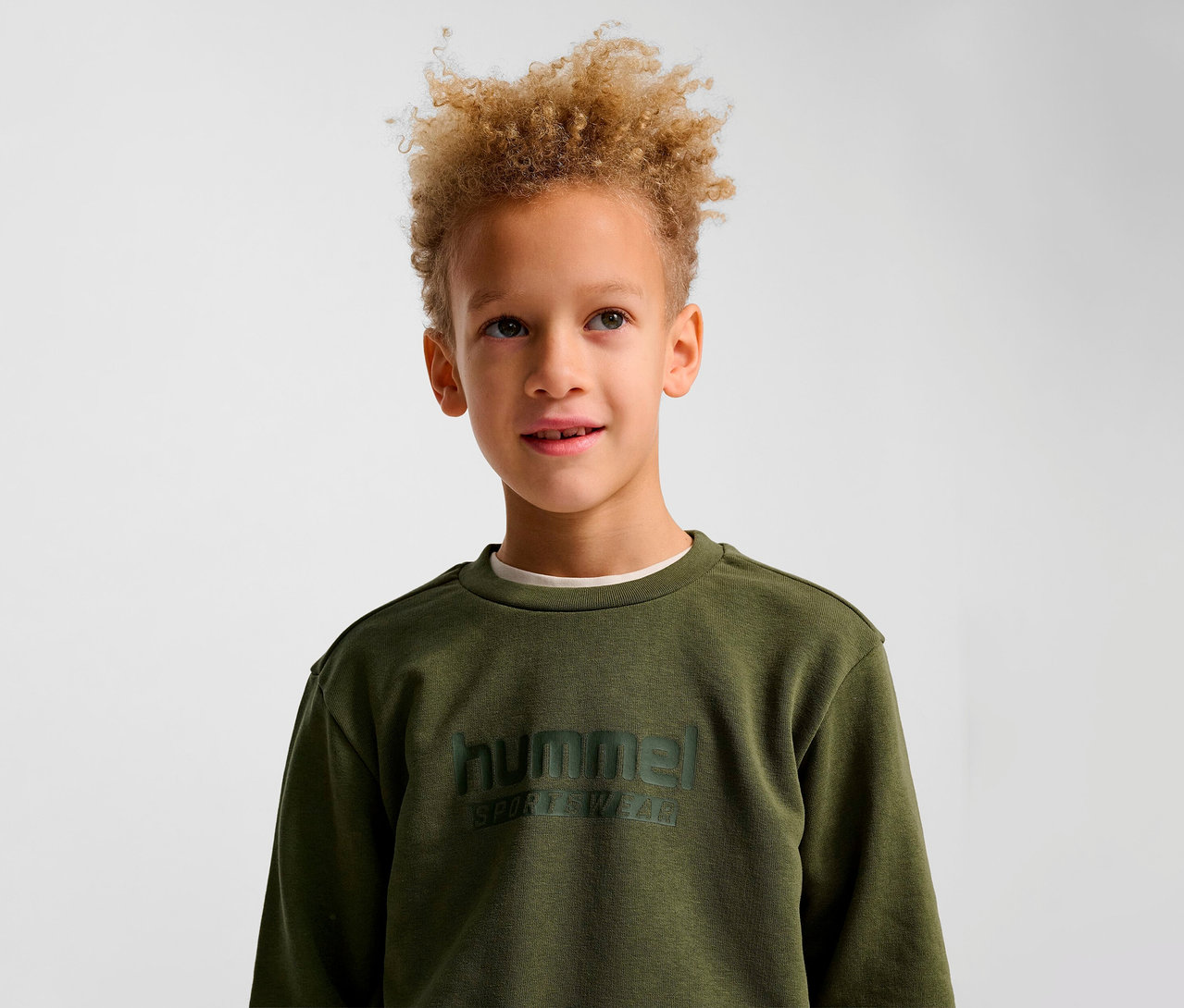 Ein Junge mit blond gelocktem Haar trägt ein grünes HUMMEL HMLJR Base-Sweatshirt.