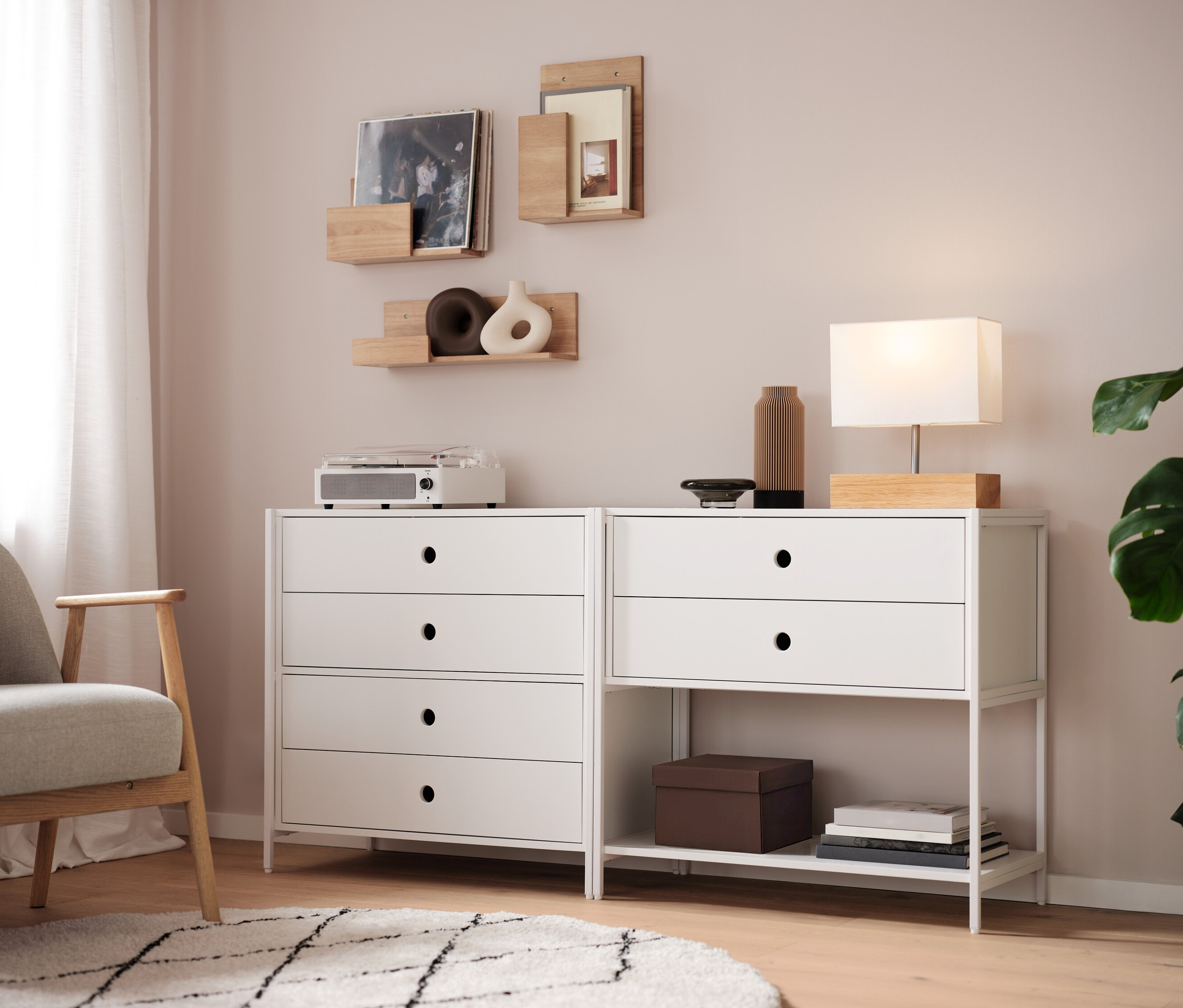 Zwei weiße Kommoden stehen nebeneinander, darauf eine Lampe, eine Vase und ein Radio. An der Wand hängen Regale.