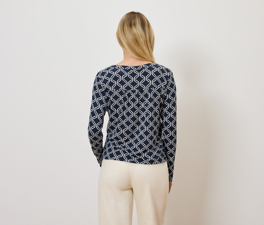 Frau mit dem Rücken zur Kamera trägt ein dunkelblaues Langarmshirt mit geometrischem Muster und eine beige Hose.