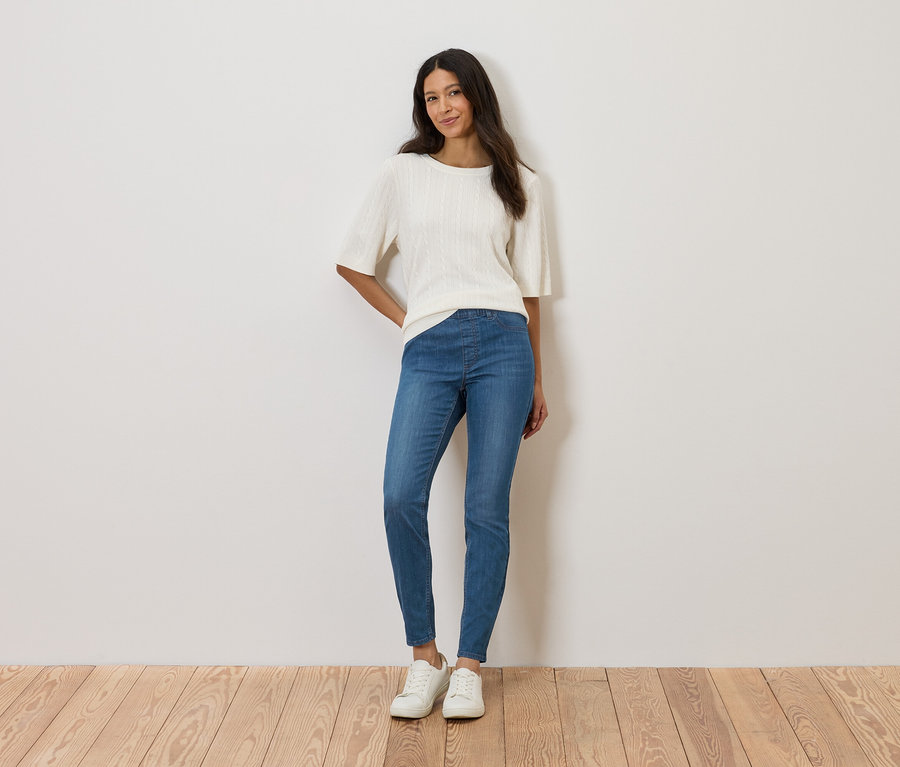 Frau steht vor einer Wand und trägt ein weißes Oberteil, blaue Jeans und weiße Schuhe.