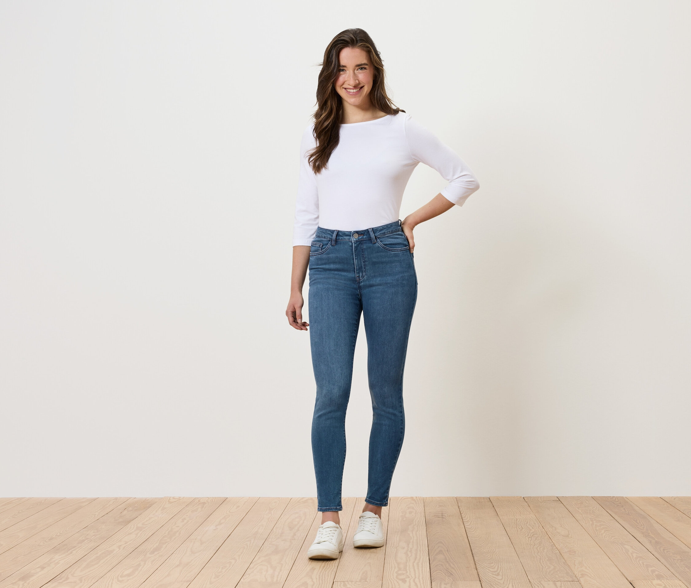 Eine Frau steht und posiert in blauen Jeans, einem weißen Oberteil und weißen Schuhen.