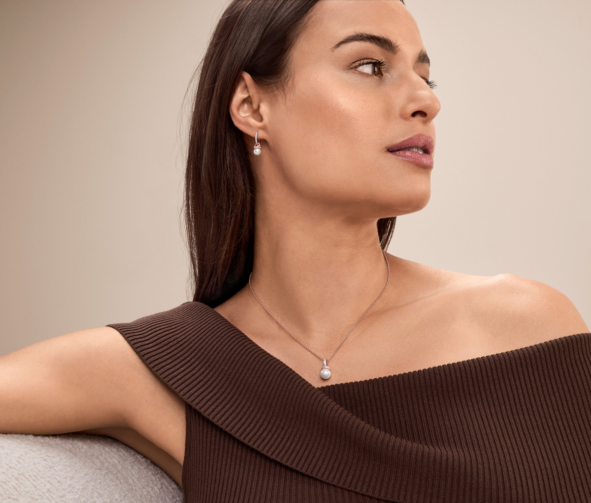 Frau mit braunen Haaren und braunem, schulterfreiem Oberteil, die eine Perlenkette und Ohrringe trägt.