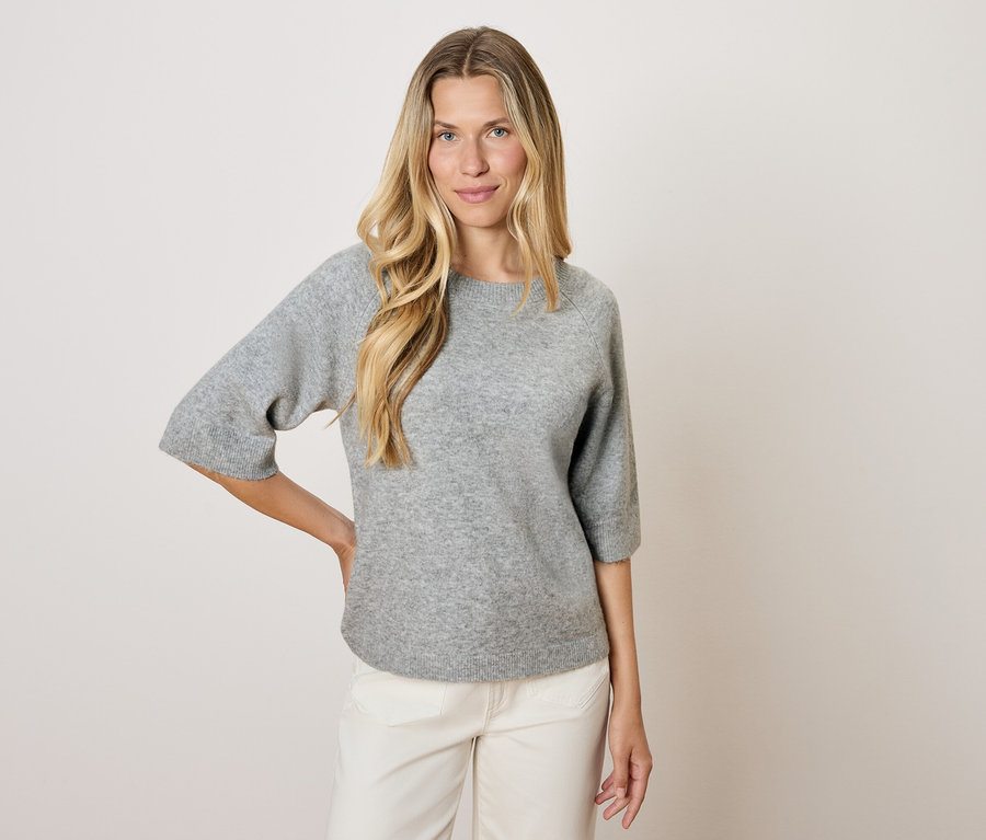 Blonde Frau in grauem Pullover mit Dreiviertelärmeln und weißen Hosen posiert vor hellem Hintergrund.