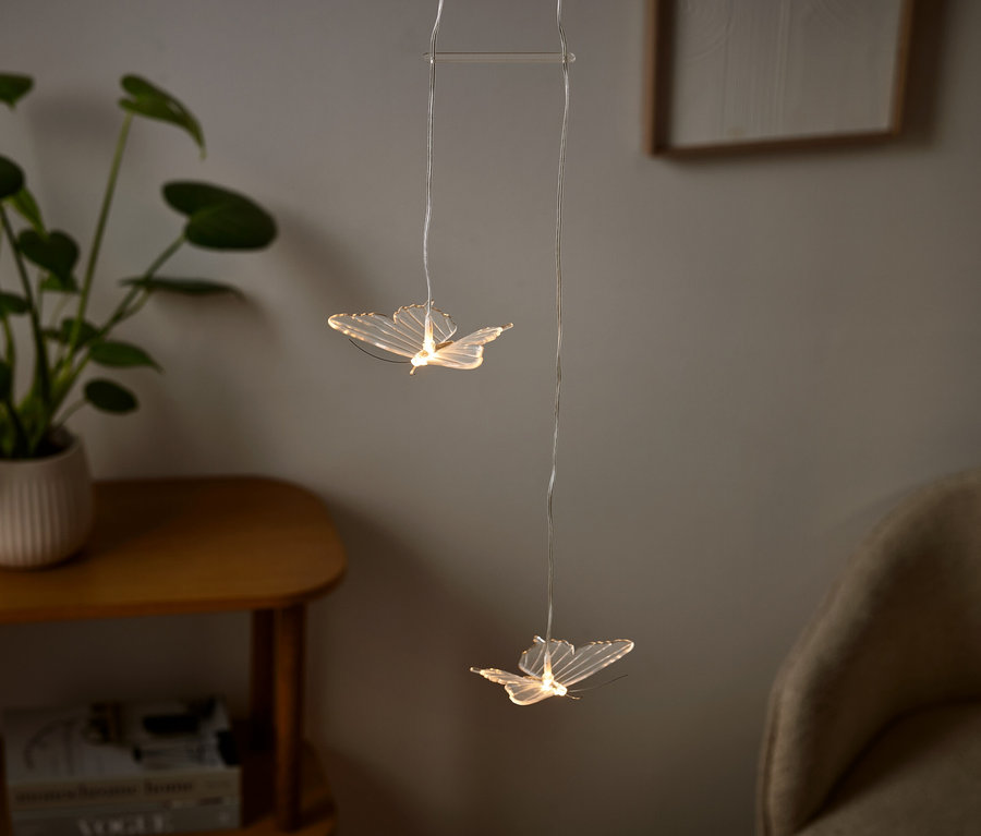 Zwei schmetterlingsförmige Lichter hängen in einem Raum, daneben eine Pflanze im Topf.