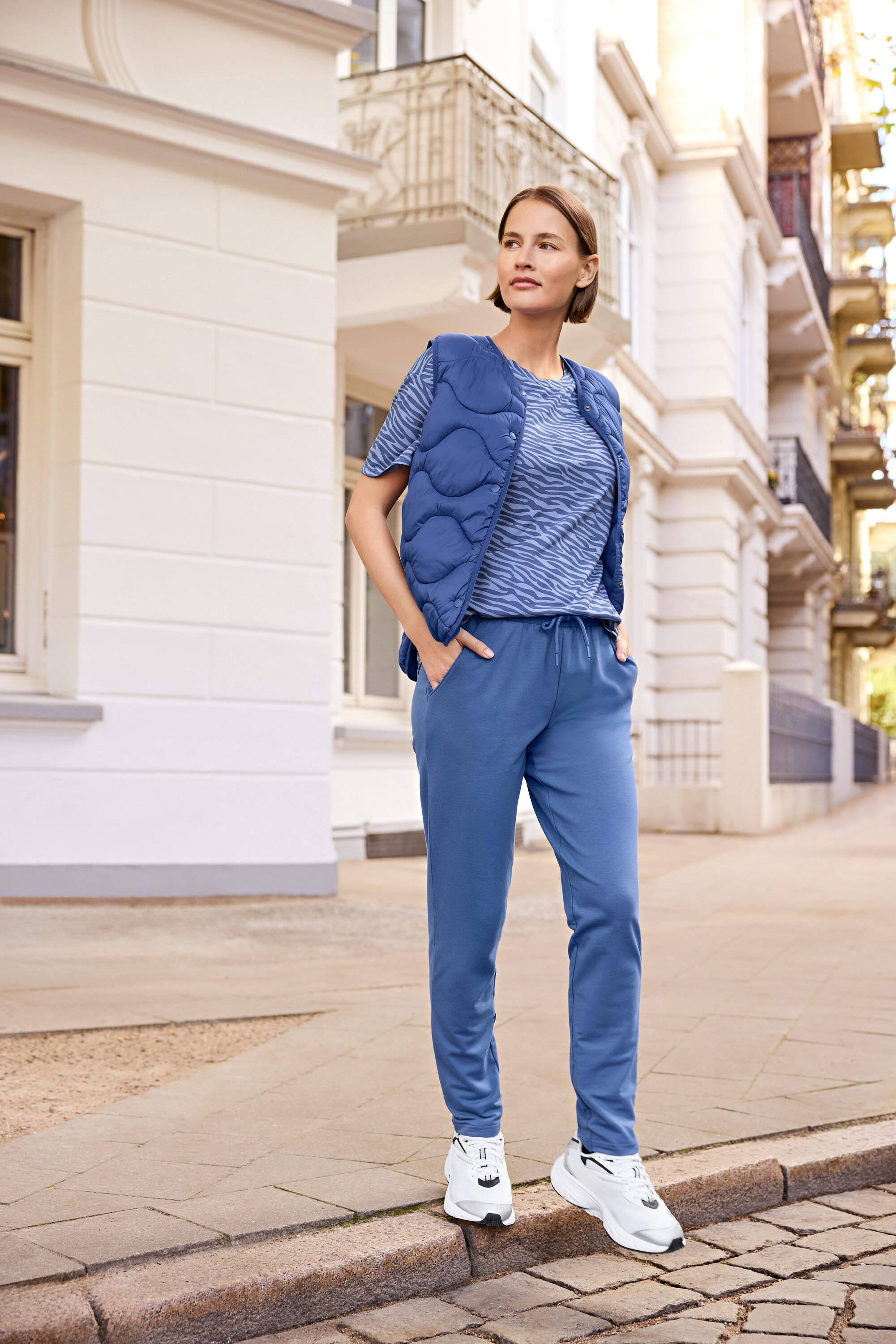 Frau steht auf dem Bürgersteig und trägt eine blaue Weste, ein blaues T-Shirt mit Zebramuster und blaue Jogginghose.