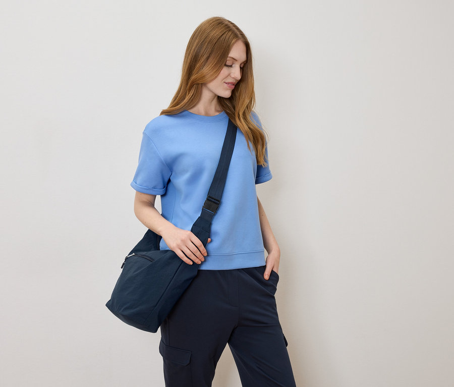 Frau mit hellbraunen Haaren, hellblauem T-Shirt und marineblauer Umhängetasche vor einem cremefarbenen Hintergrund.