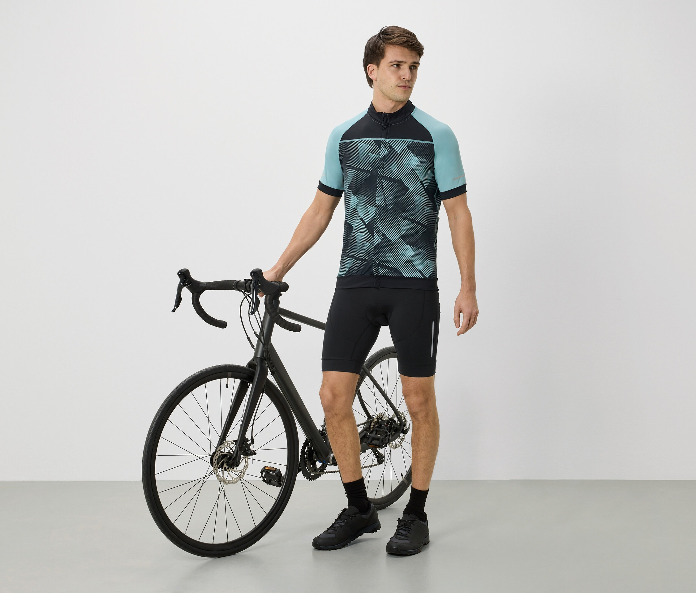 Mann steht neben einem schwarzen Fahrrad, trägt ein blau-schwarzes Trikot, schwarze Shorts und schwarze Schuhe.