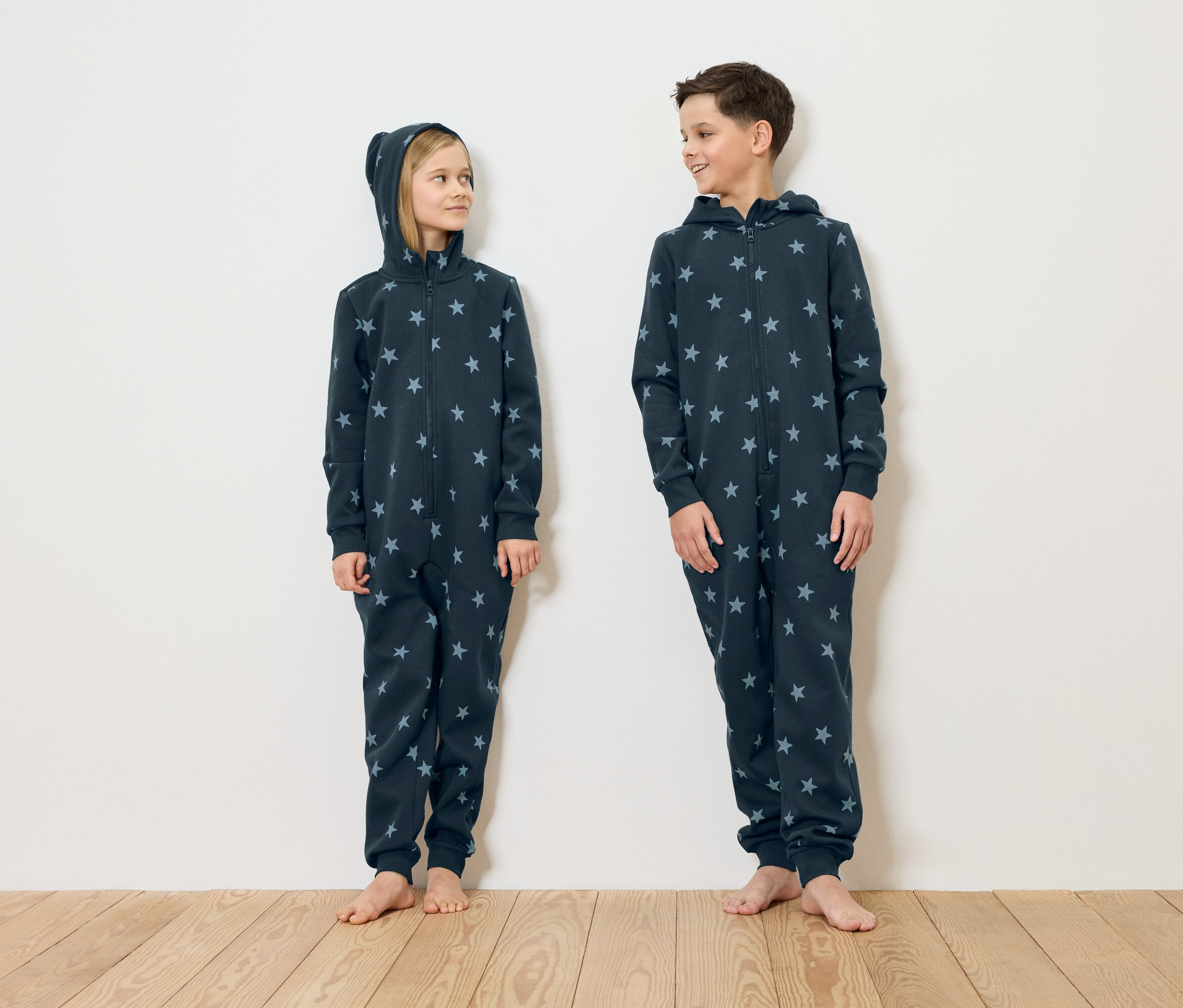 Ein Mädchen und ein Junge stehen barfuß vor einer weißen Wand und tragen marineblaue Schlafanzüge mit Sternenmuster.