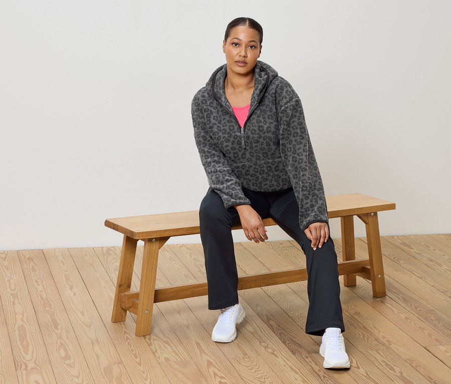 Frau sitzt auf einer Holzbank und trägt einen grauen Fleece-Pullover mit Leopardenmuster, eine pinkes Top, eine dunkle Hose und weiße Turnschuhe.