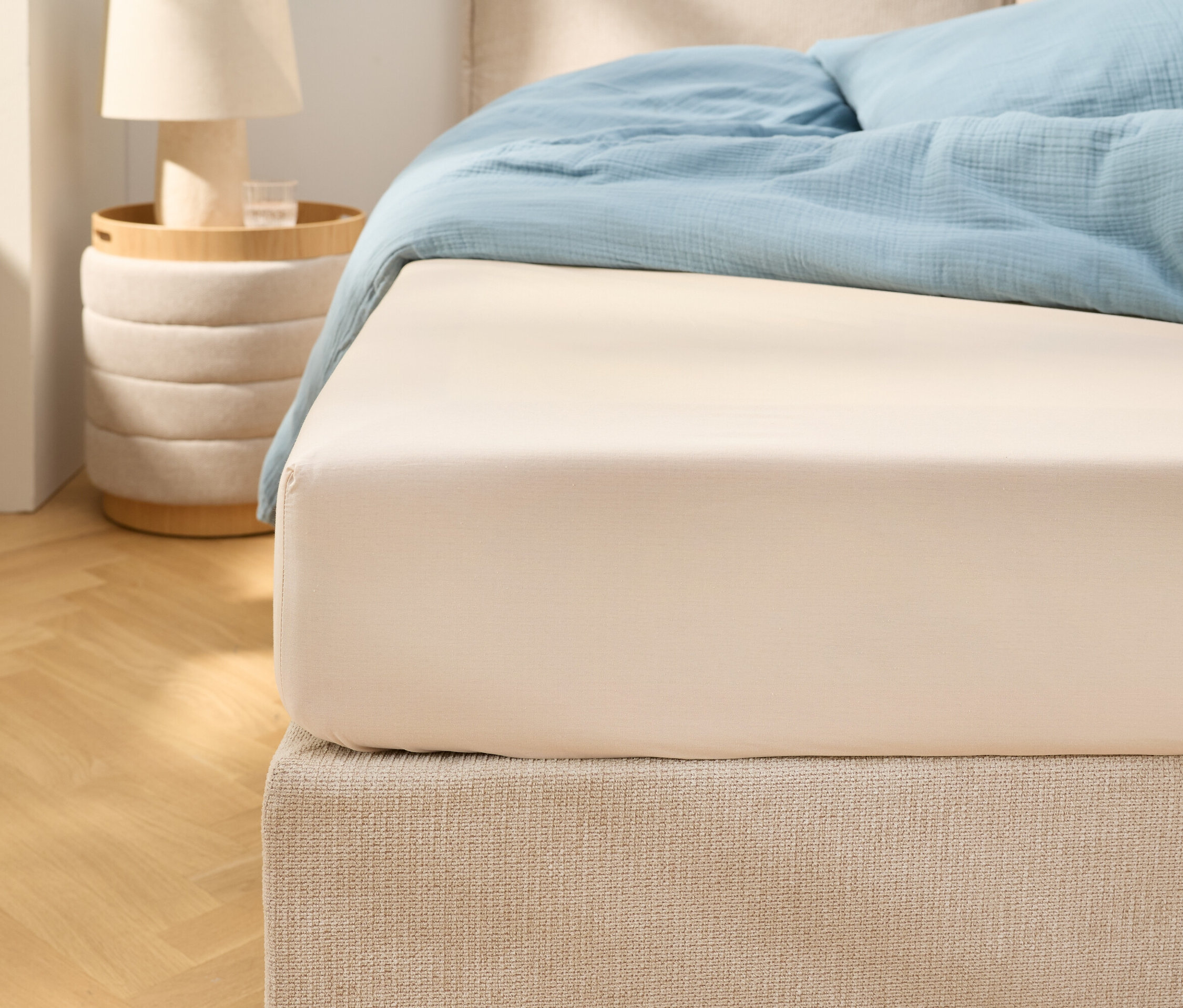 Nahaufnahme eines beigen Spannbettlakens auf einem Bett mit hellblauer Bettwäsche. Ein Nachttisch mit einer Lampe steht daneben.