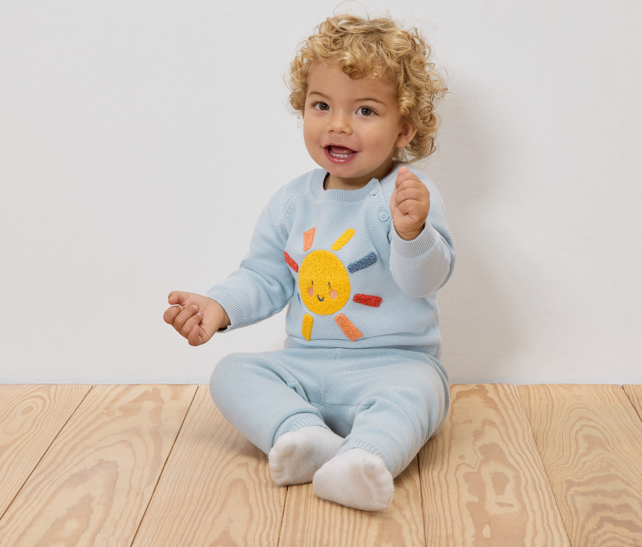 Ein lächelndes Baby mit blonden Locken sitzt auf einem Holzboden und trägt einen hellblauen Pullover mit einer aufgestickten Sonne und eine passende Hose.