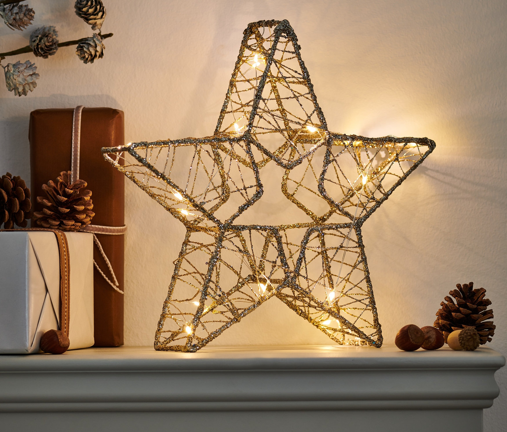 Ein LED-Stern aus Draht steht auf einem Sims neben Geschenken und Tannenzapfen.