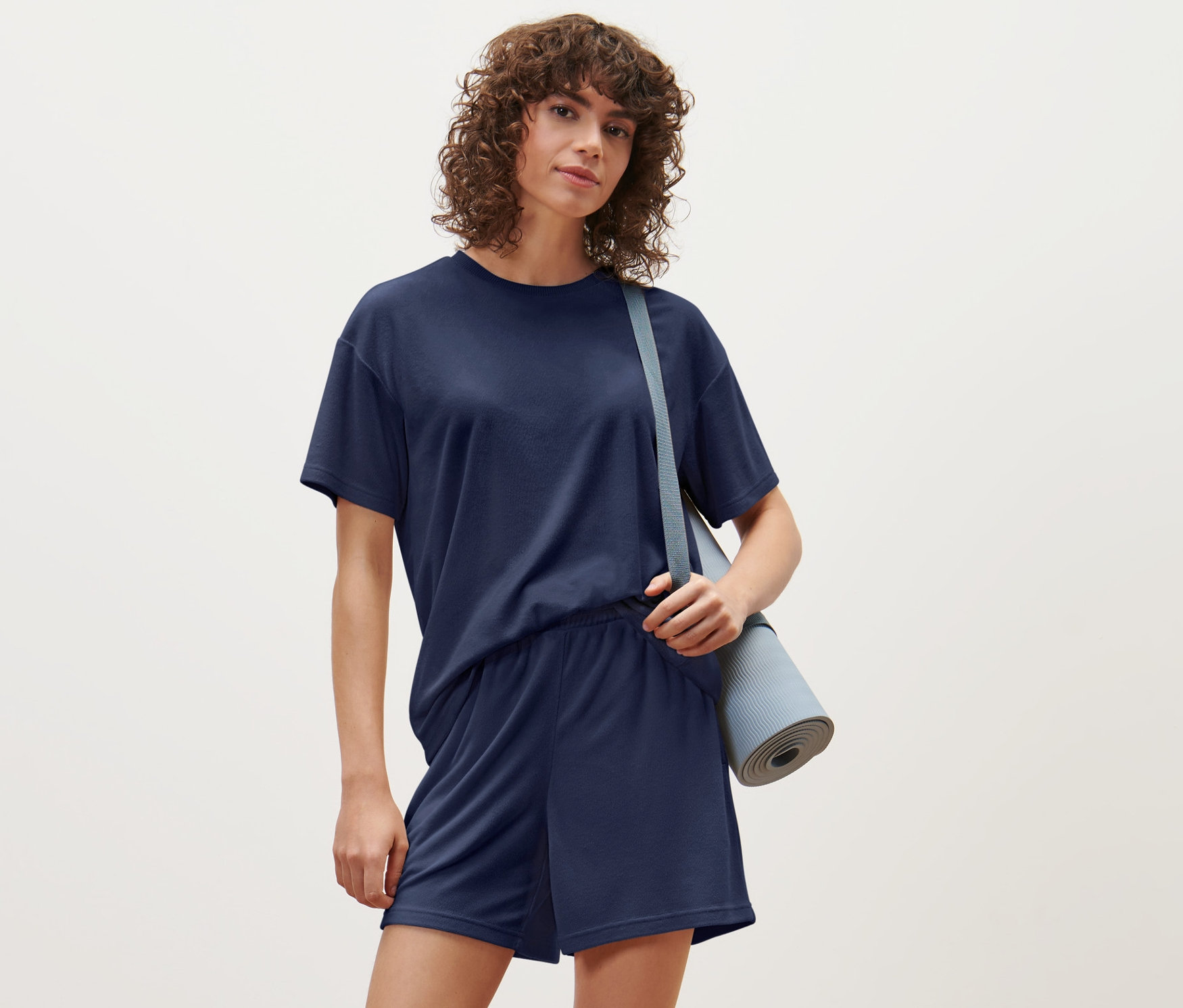 Frau trägt ein Frottee-Sportshirt und Shorts, mit einer Yoga-Matte über der Schulter.