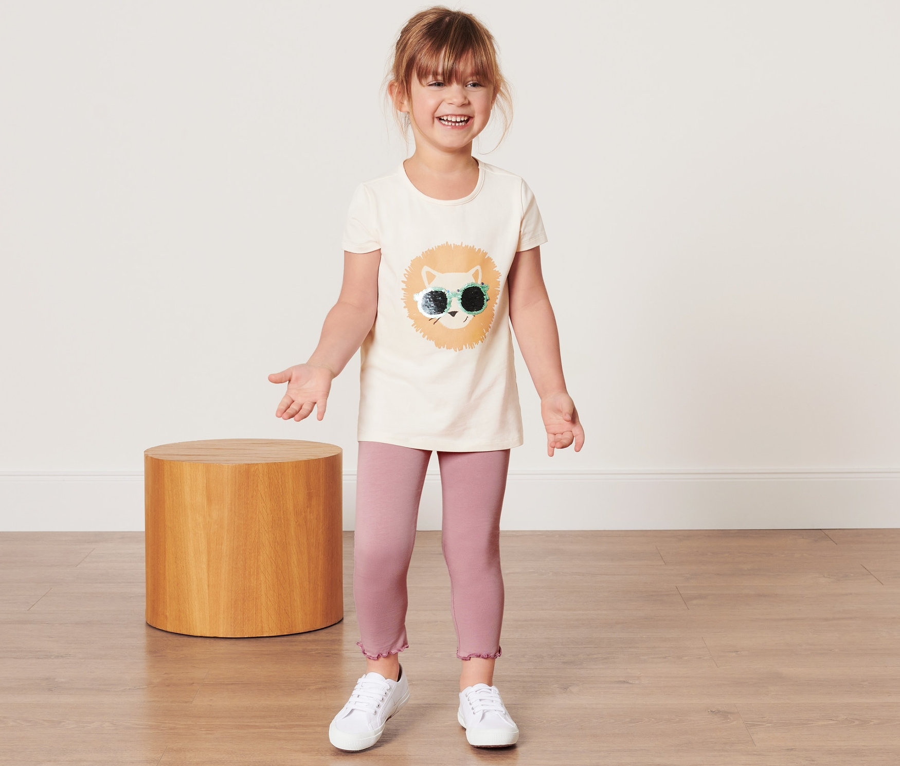 Ein lächelndes Mädchen steht und trägt Kinder-Leggings und ein T-Shirt mit einem Löwenaufdruck.