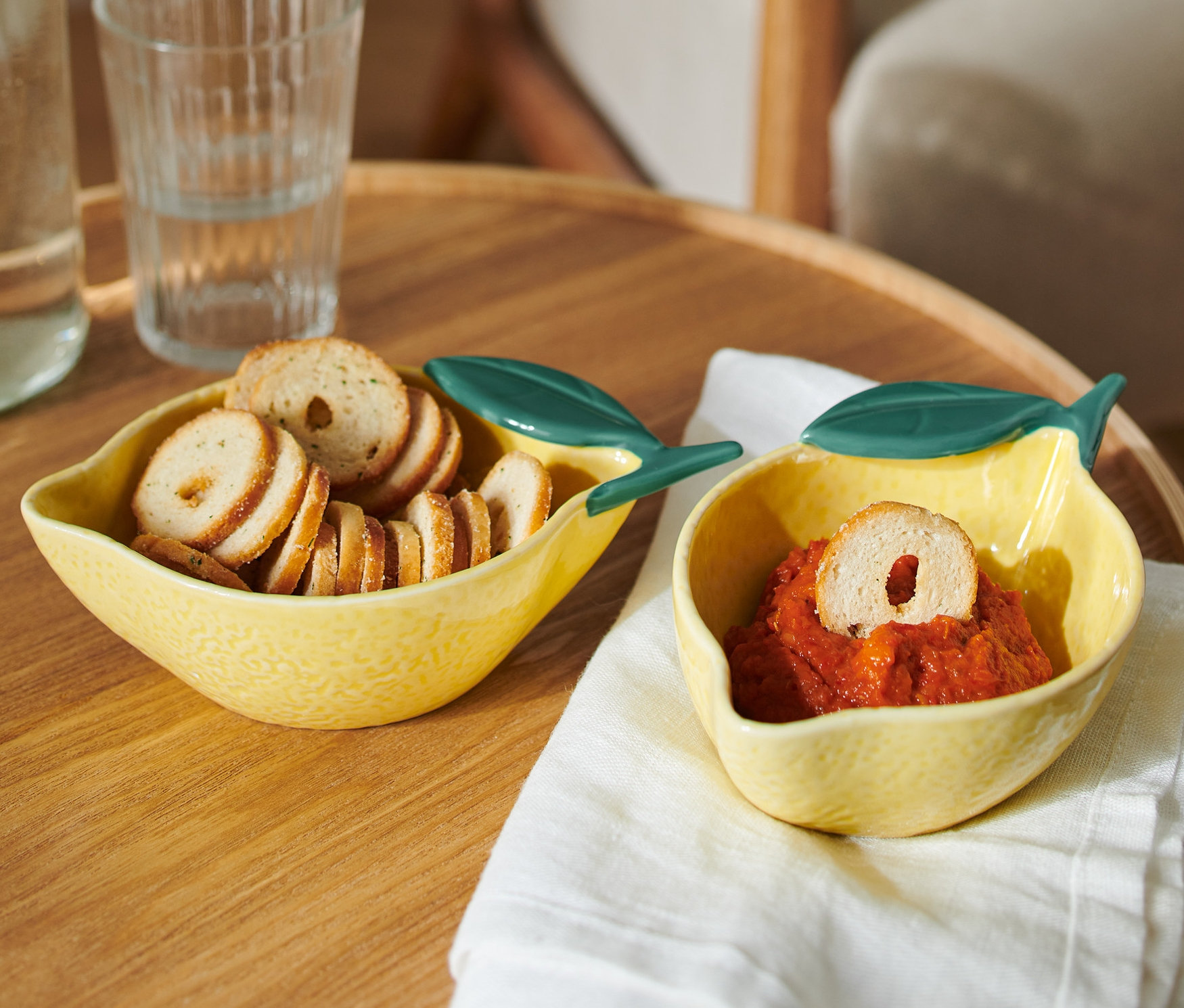 Zwei zitronenförmige, gelbe 2 kleine Schalen stehen auf einem Tisch. Eine ist mit Brotscheiben, die andere mit einem roten Dip gefüllt.