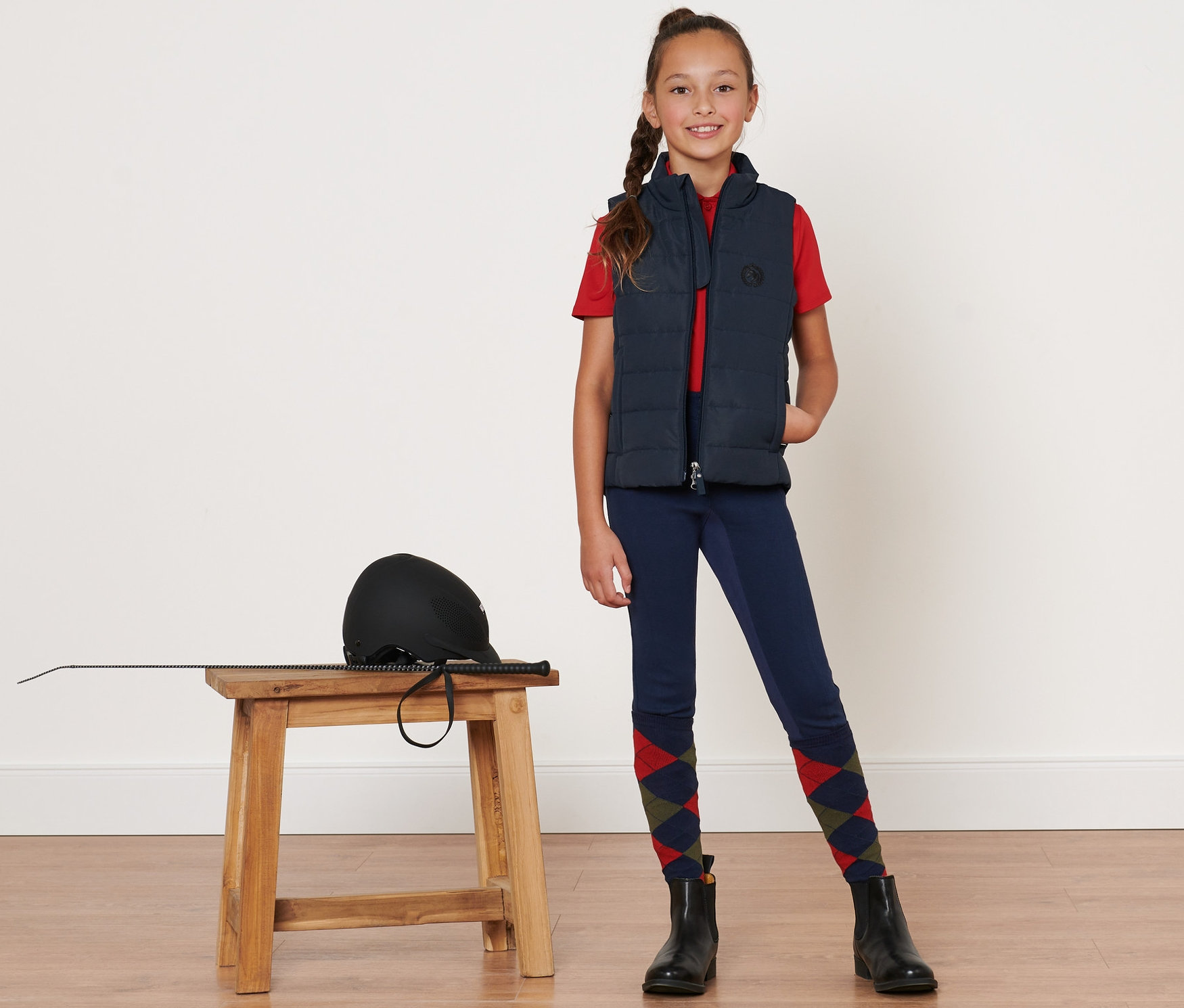 Mädchen in Kinder-Reitweste steht neben Hocker mit Helm und Peitsche.